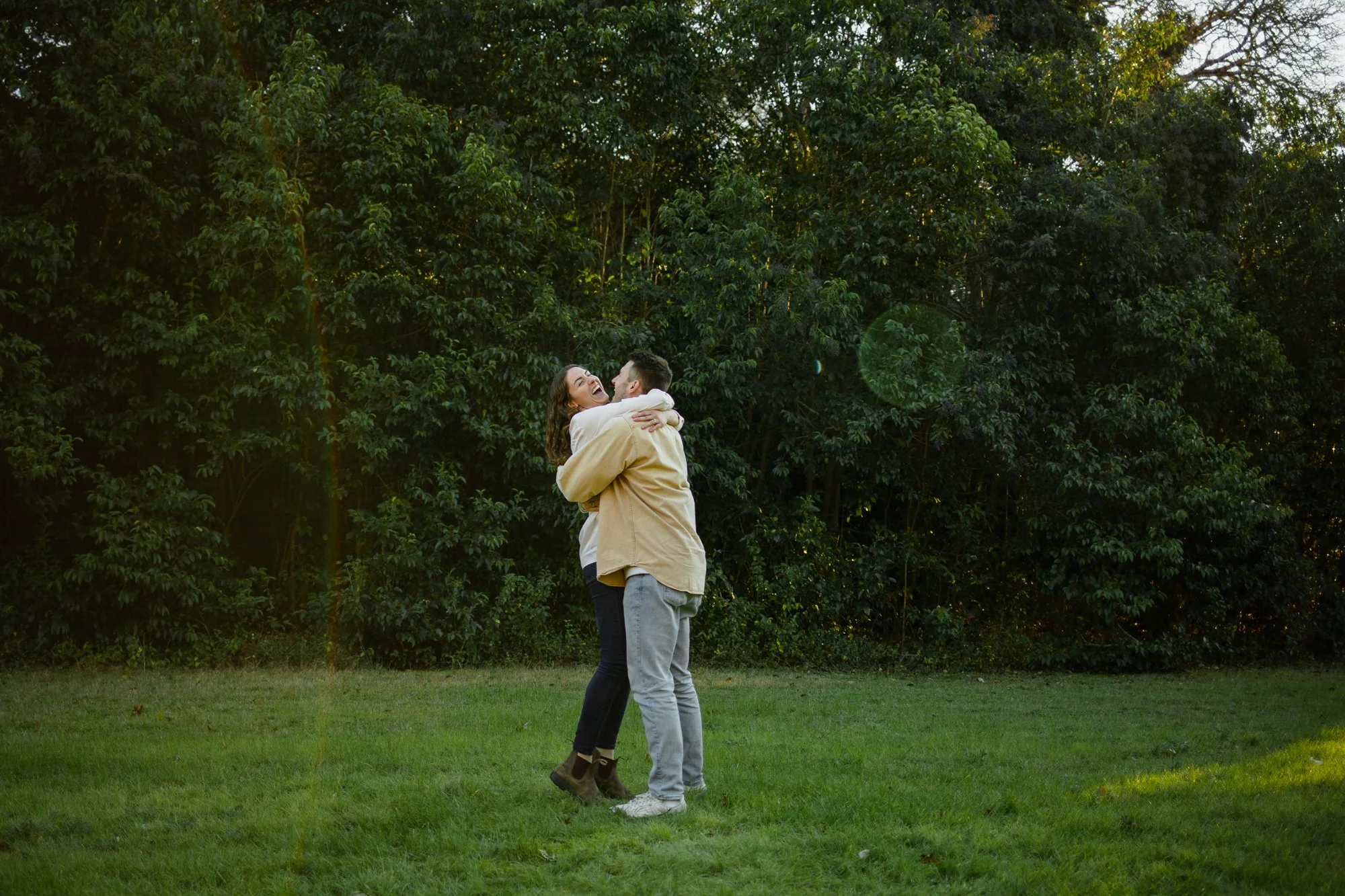A couple hugging and laughing outdoors on a grassy field with trees in the background, during sunset.