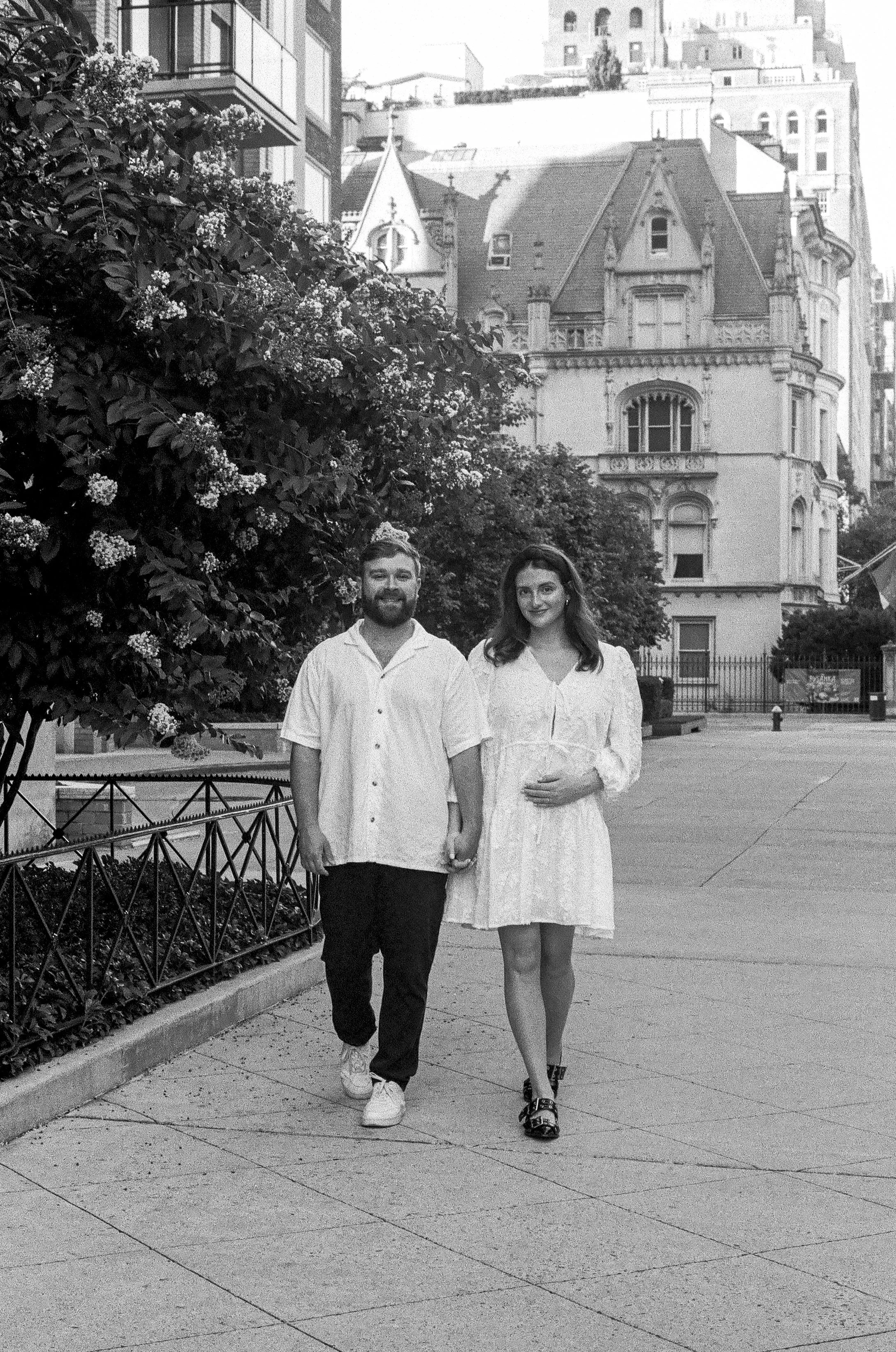 A black and white photo of a man and a pregnant woman walking hand in hand outdoors in an urban setting. The man has a beard, glasses, and is wearing a short-sleeved button-up shirt, dark pants, and sneakers. The woman has long, wavy hair and is wear