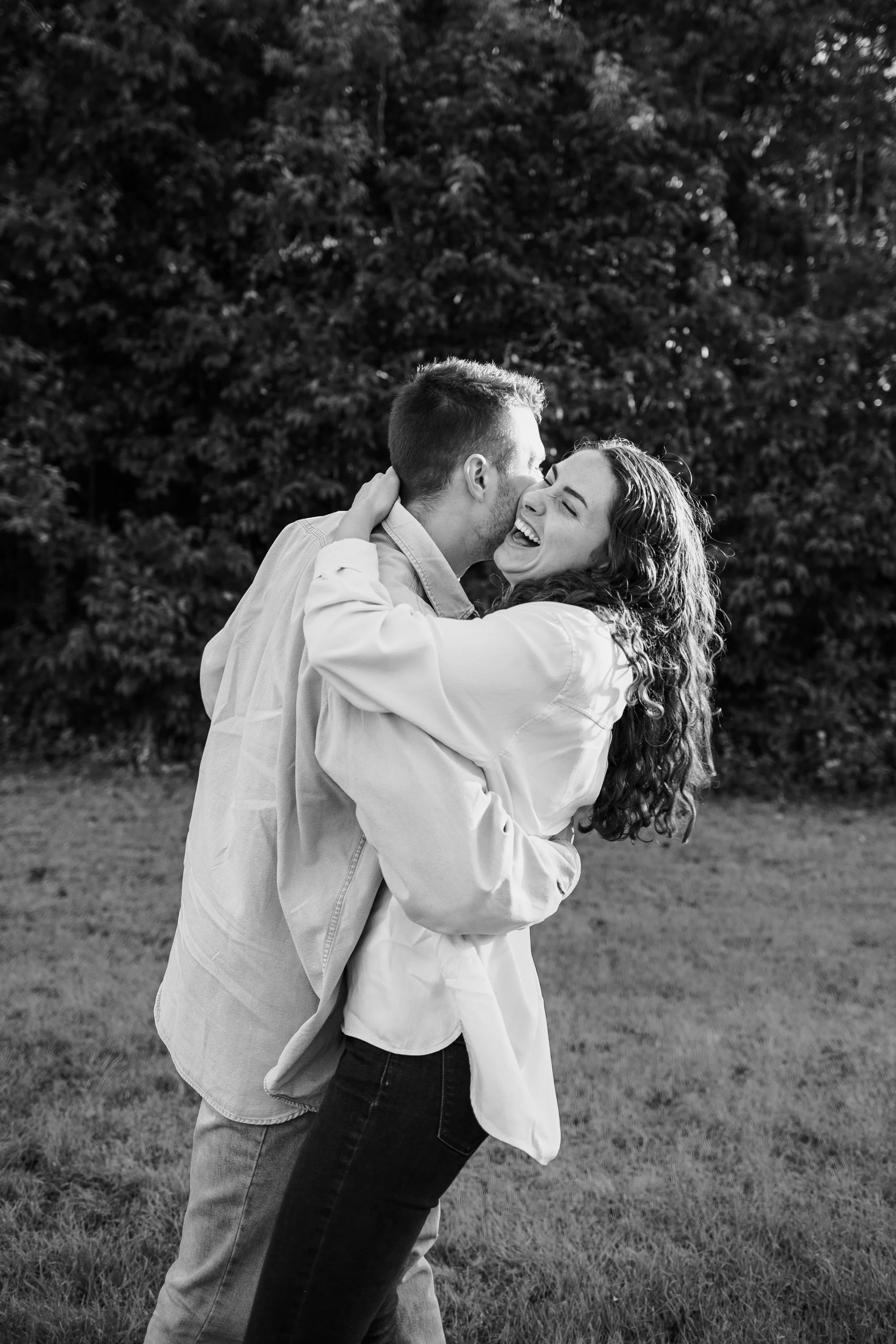 A joyful couple, a man and woman, sharing a kiss outdoors on a grassy area with trees in the background, smiling and embracing each other.