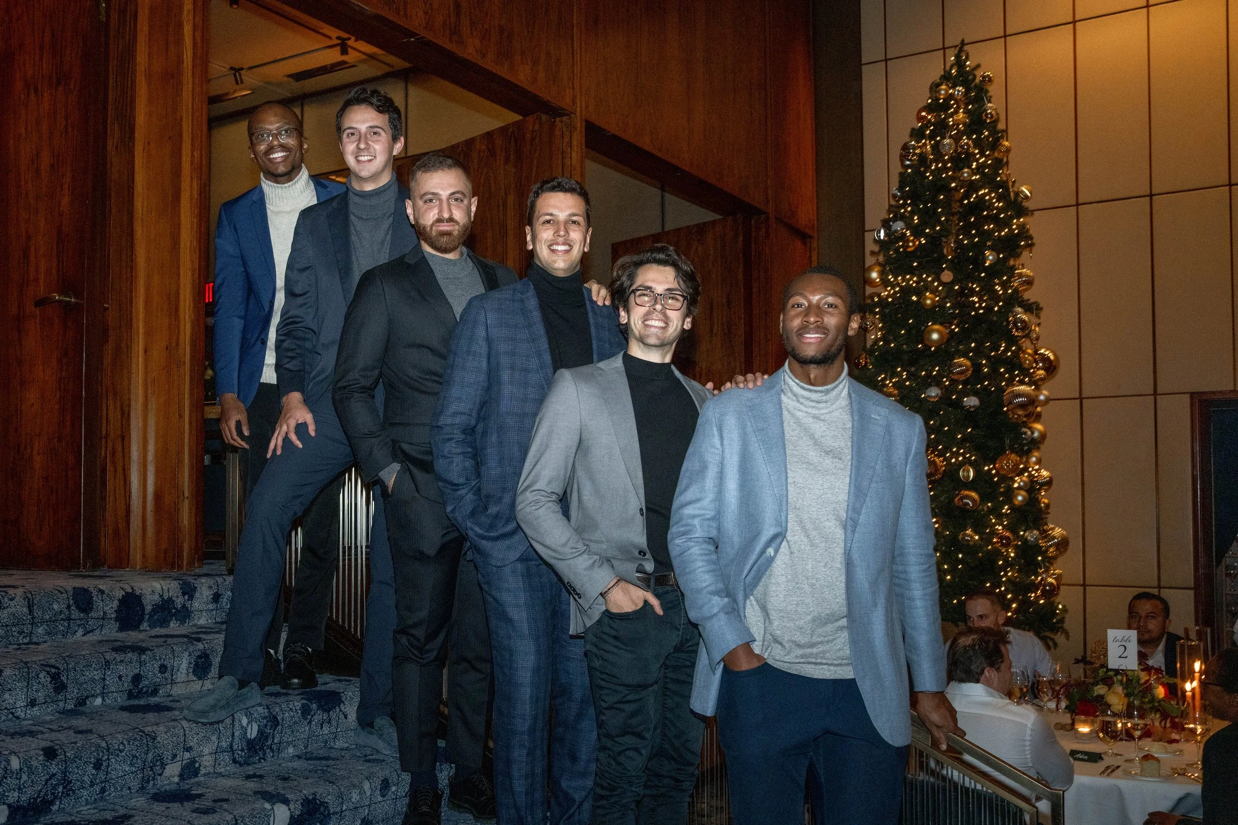 Group of six men dressed in suits and smart casual attire standing with a decorated Christmas tree in the background.