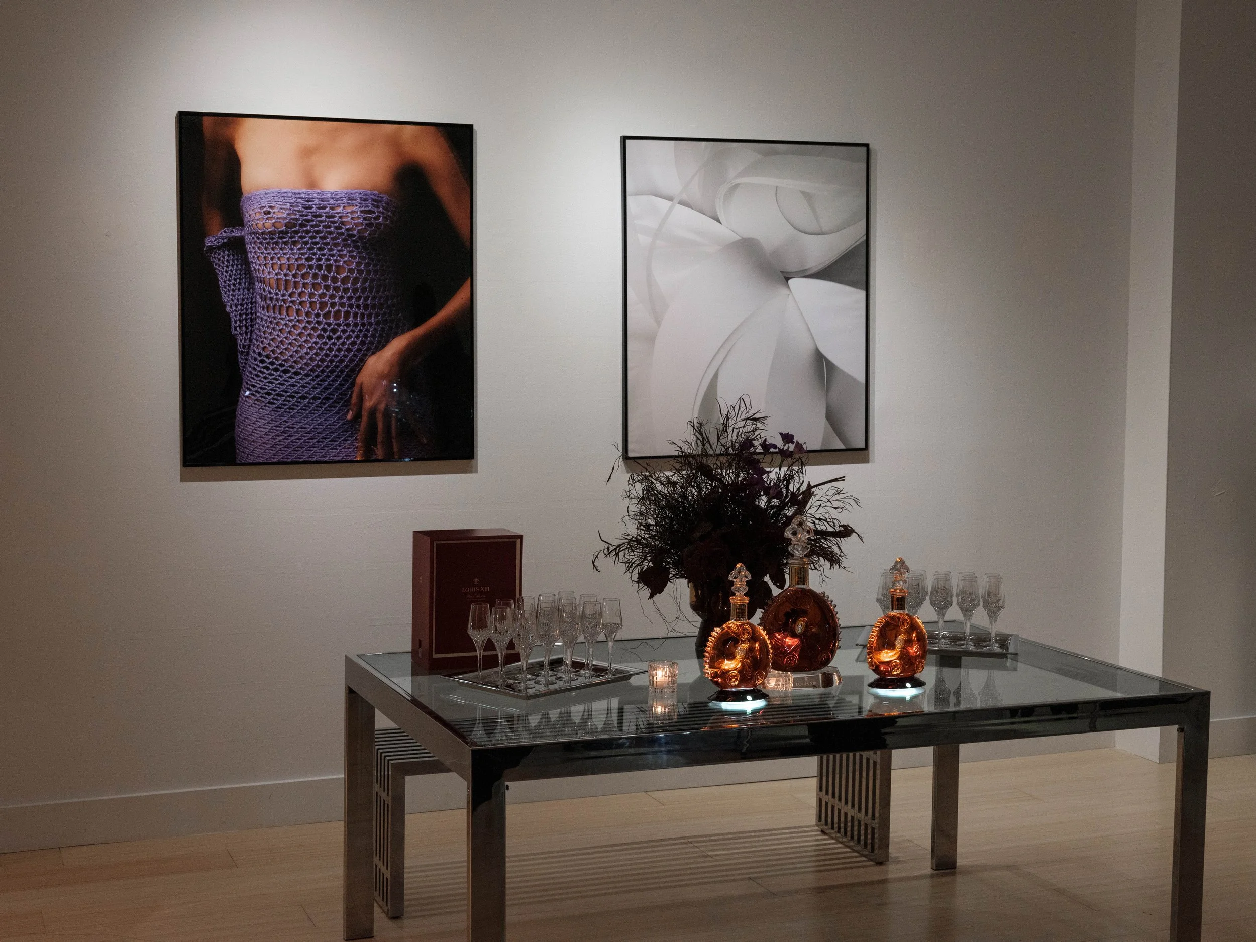 An art gallery scene features two large framed photographs on a white wall; one of a person wearing a purple crochet dress, and the other of a white floral sculpture. A glass table below holds three orange bottles, multiple empty champagne glasses, a