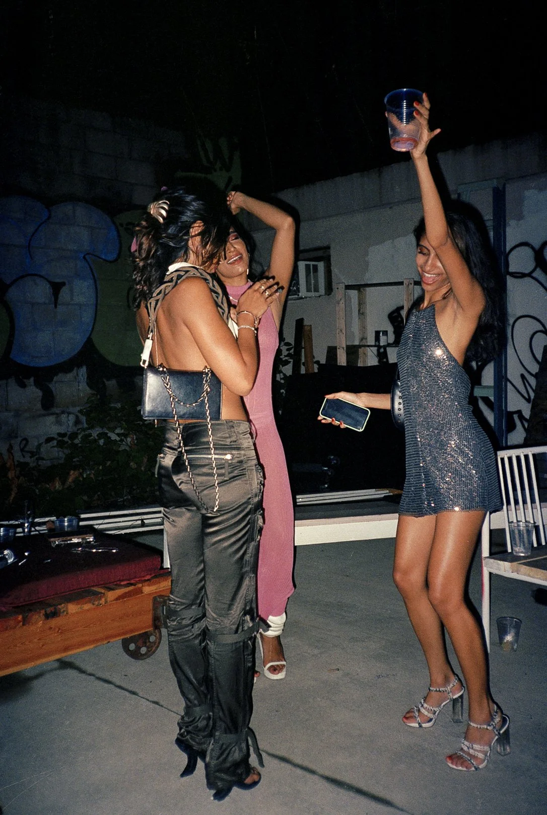 Three women dancing and having fun at a party, with one woman holding a cup and another holding a phone, at night