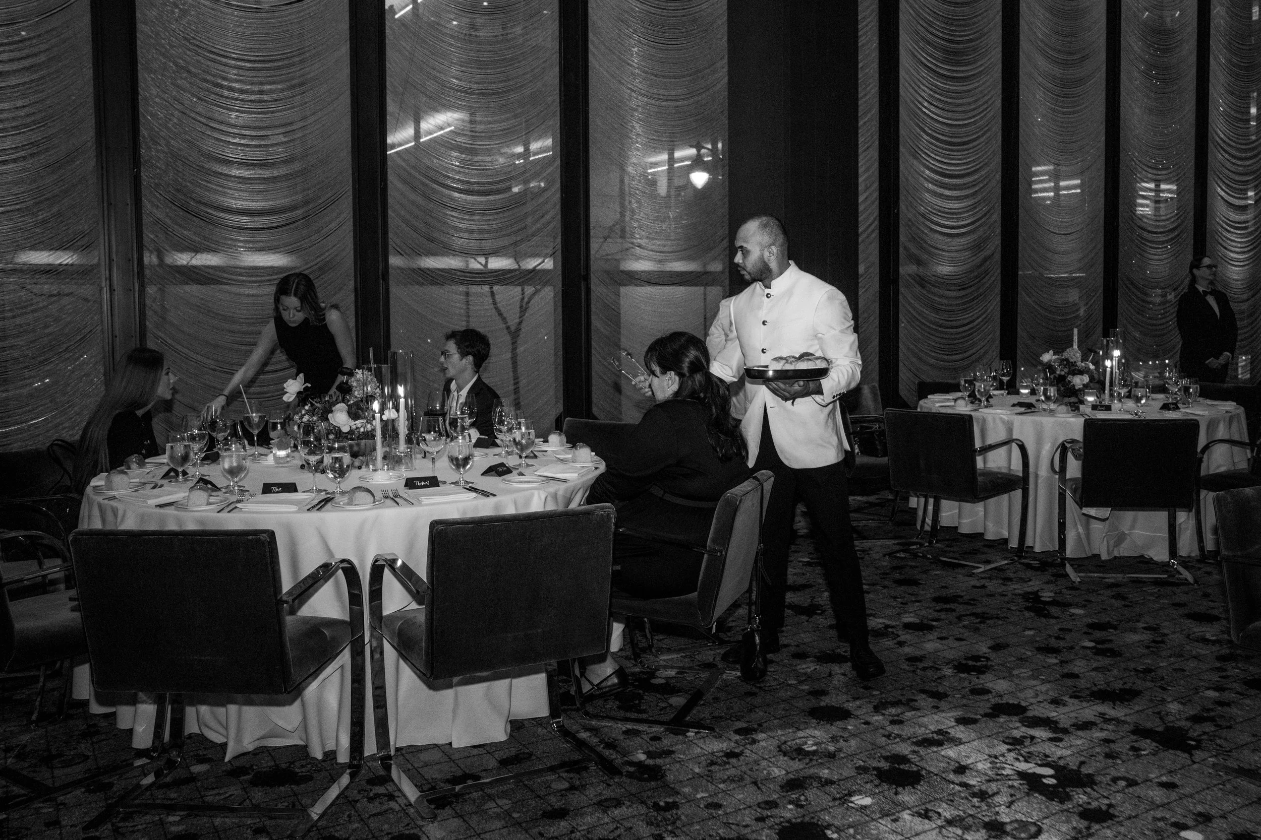 Black and white photo of a formal dining event with waiters serving guests seated at round tables with floral centerpieces.