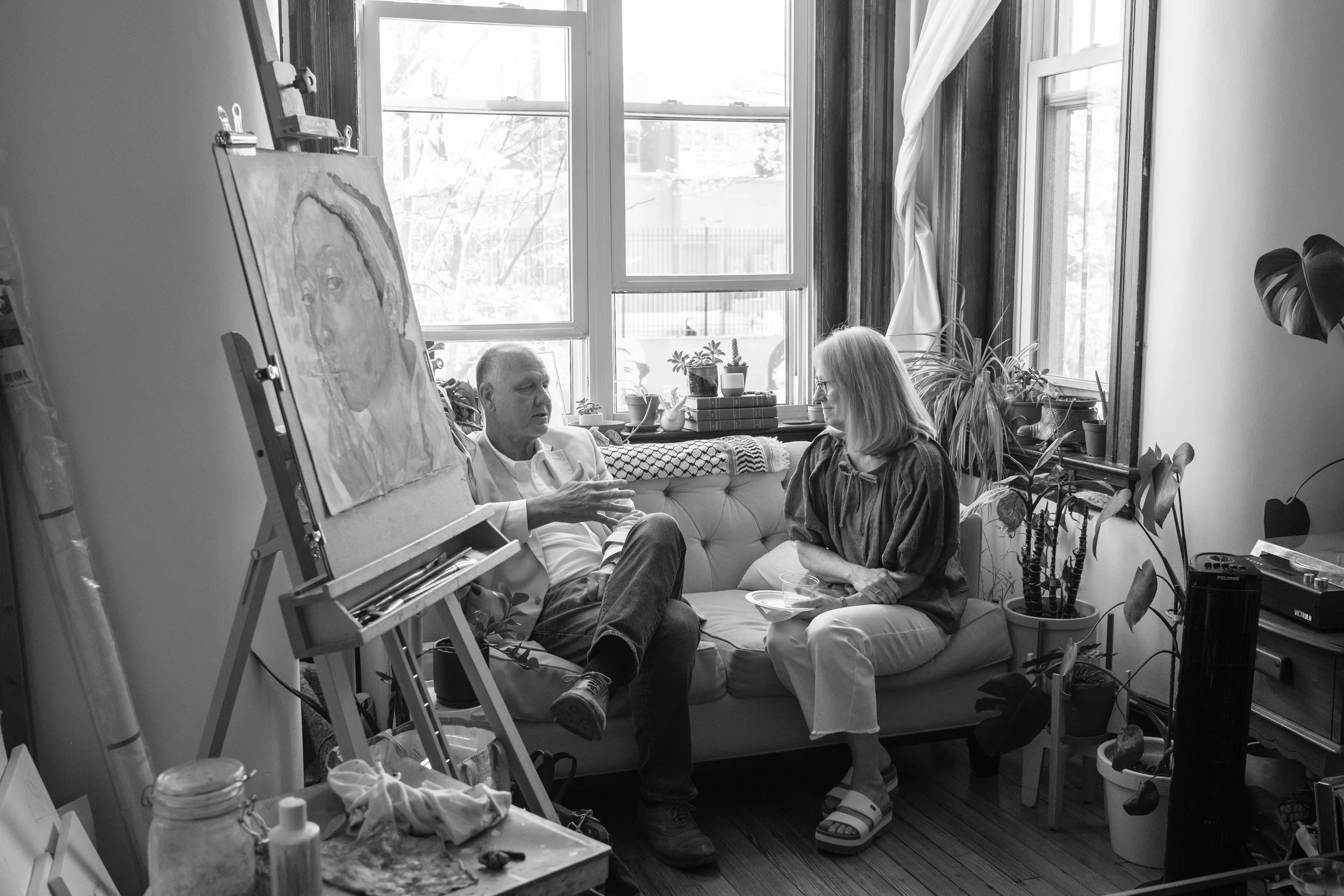 A black and white photo of two people sitting on a couch in a bright room. The man is gesturing with his hand while talking, and the woman listens. The room has large windows with plants and objects on the windowsills, and an easel with a painting.