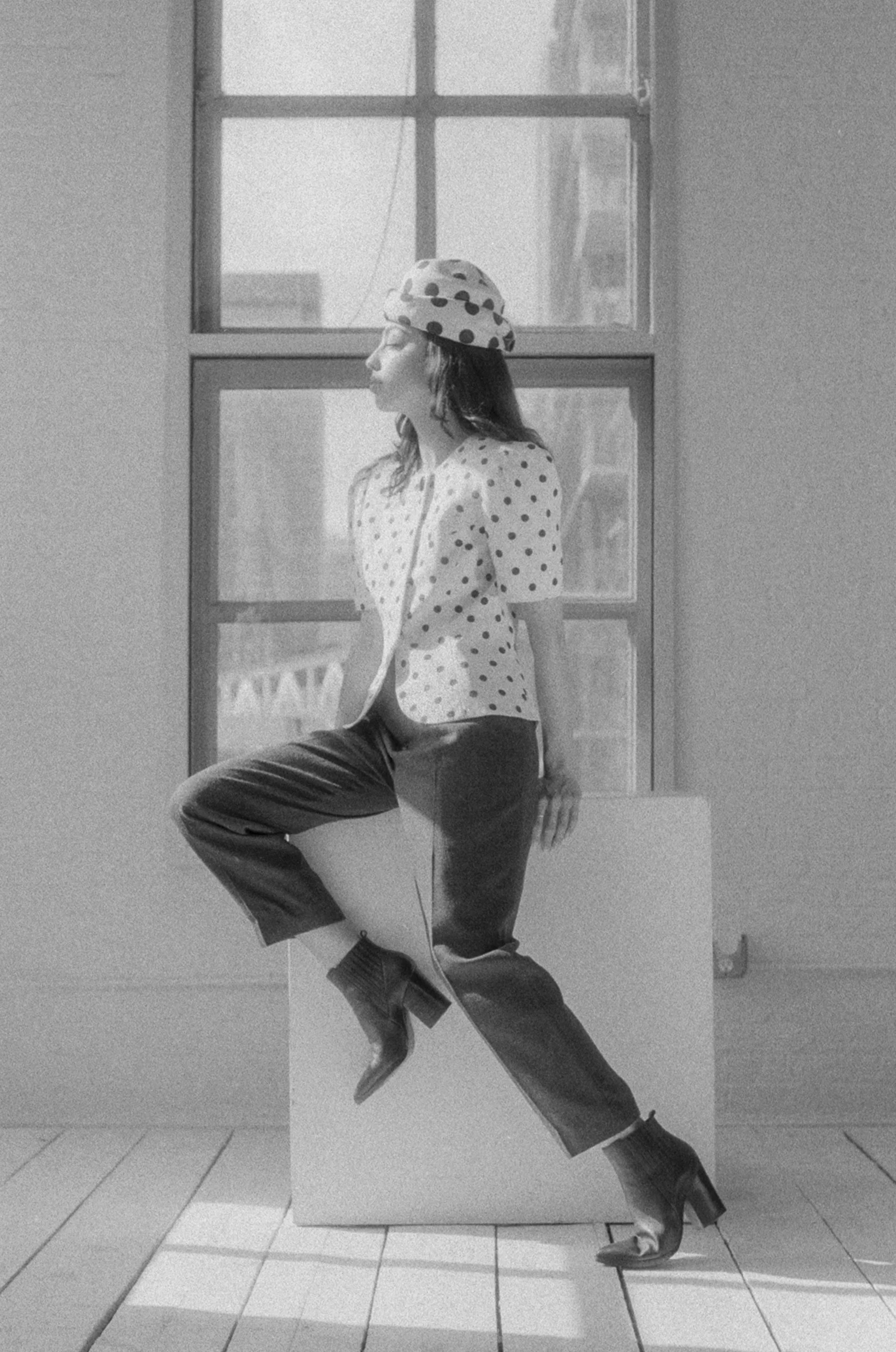 Black and white photo of a woman sitting on a white block by a window, wearing a polka dot hat, polka dot blazer, pants, and high-heeled boots, looking to the left.