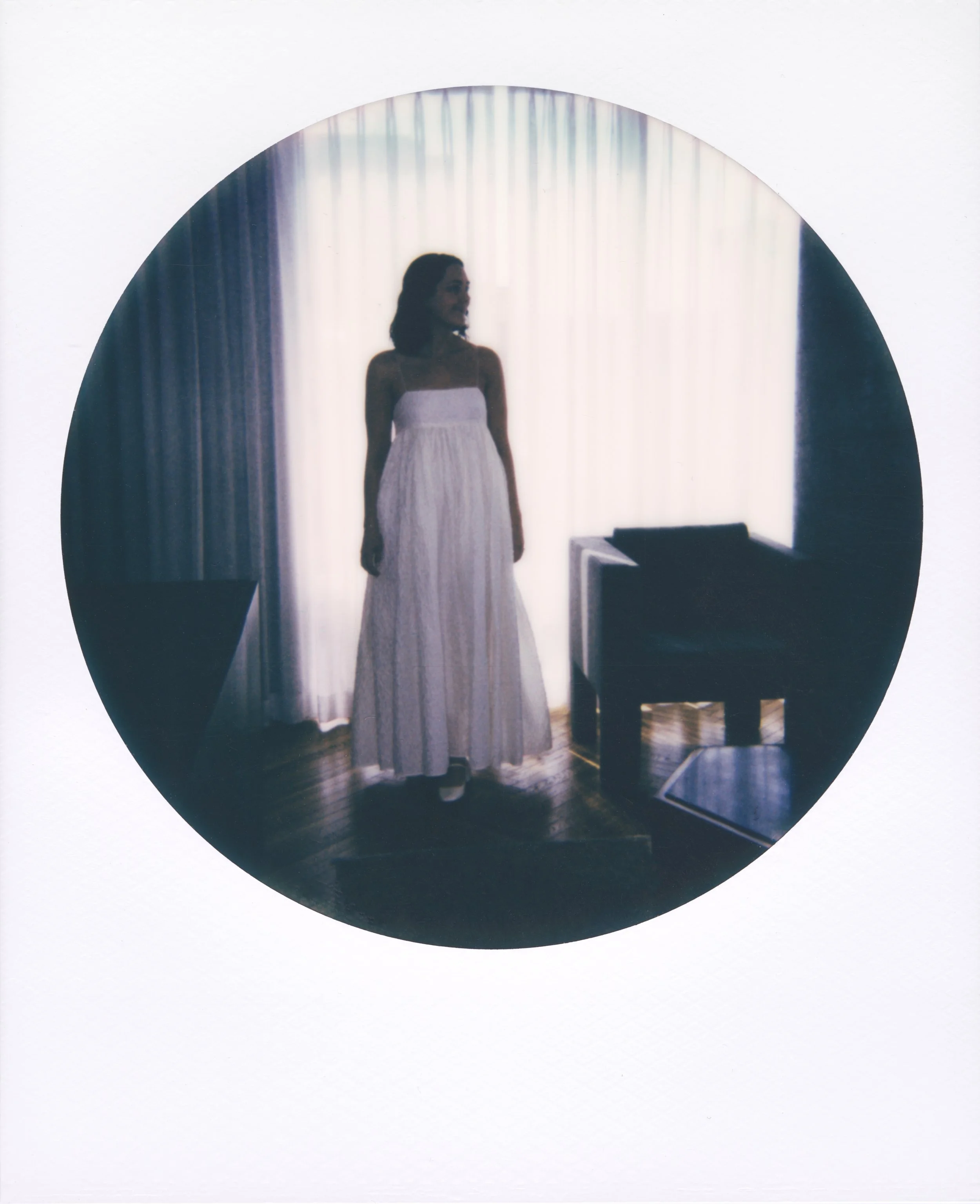 Round-frame polaroid of a woman standing in front of a backlit window, wearing a white strapless dress, with curtains, a table, and a chair in the background.