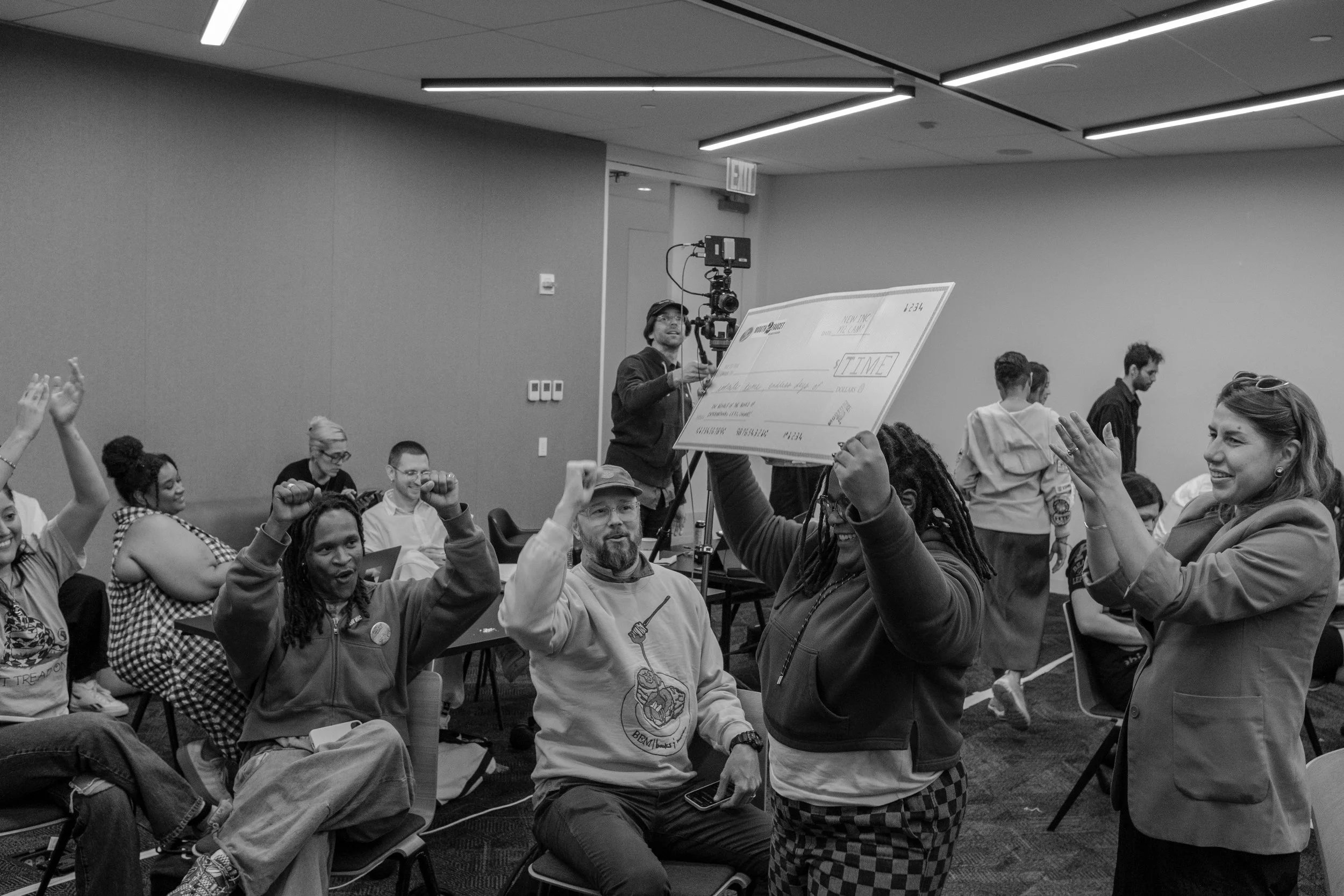 People celebrating and cheering in a large room, with one person holding a large check, others raising their fists or hands, and a camera operator recording the scene.