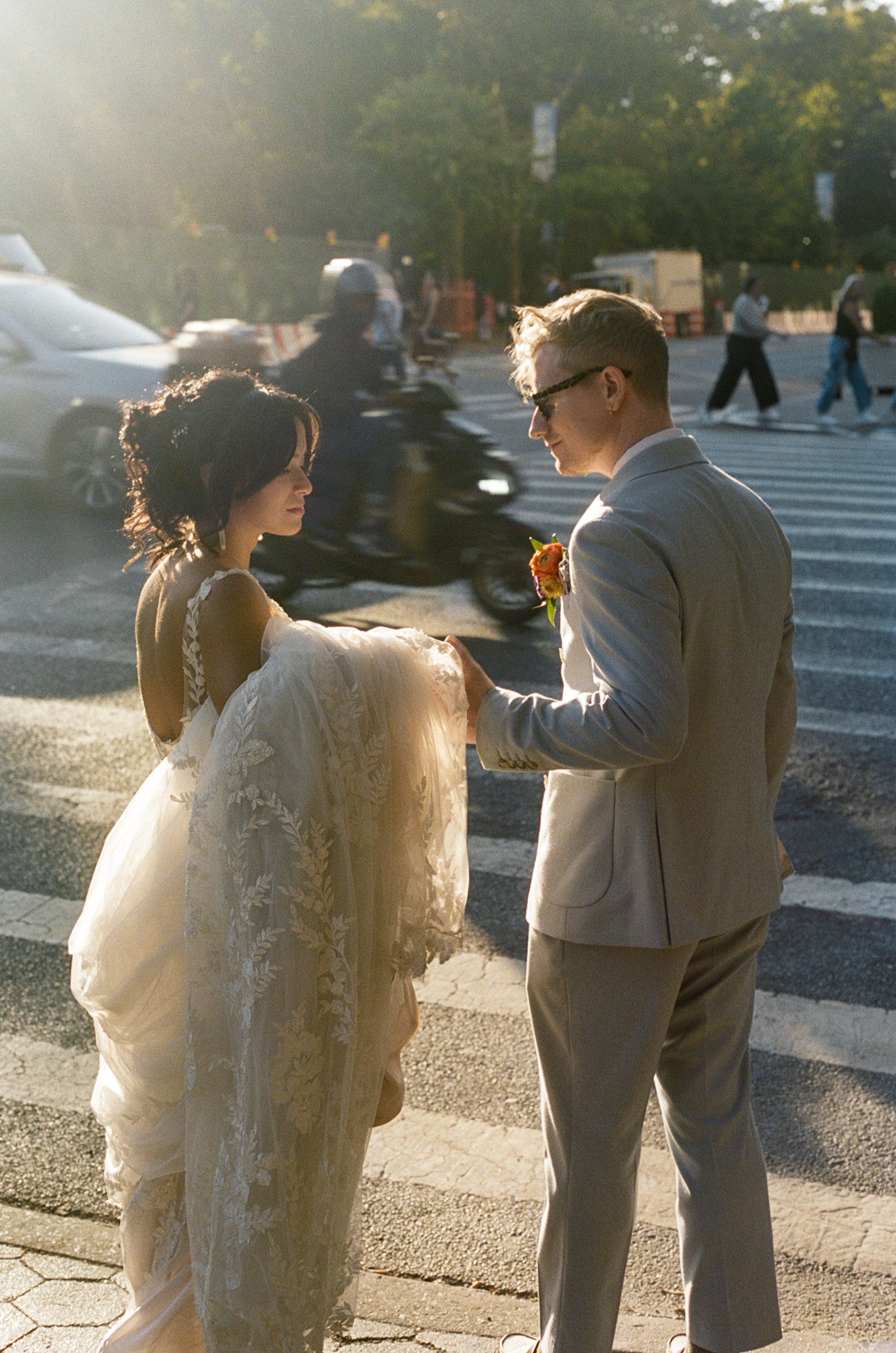 A bride and groom on a city street at sunset, with pedestrians and vehicles in the background. The groom is holding the bride's gown.