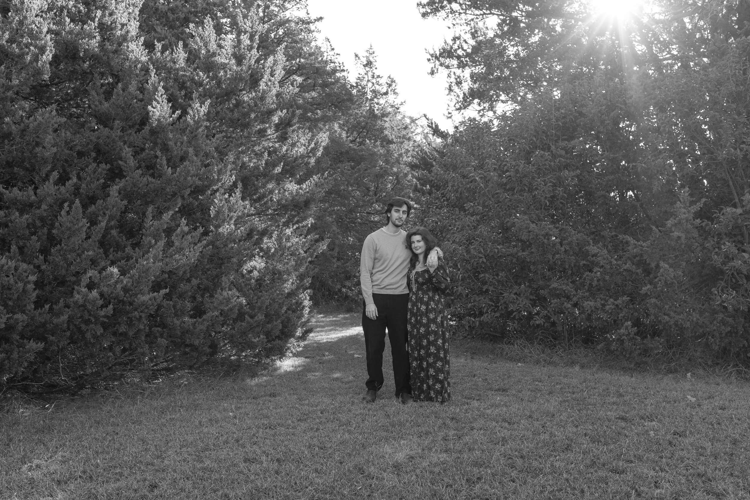 A man and woman standing close together outdoors surrounded by trees and bushes, with sunlight shining from the top right corner.