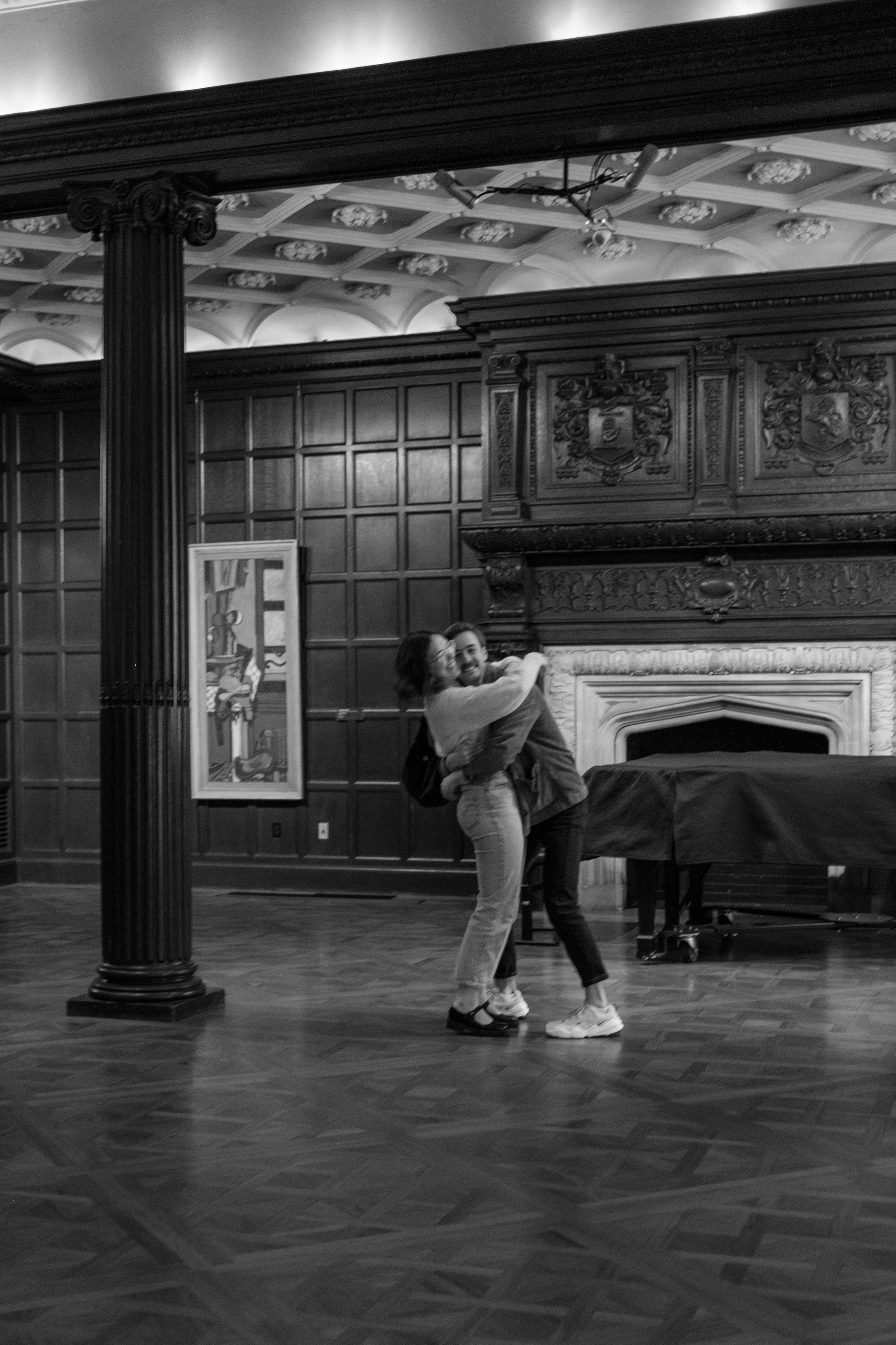 A couple hugging and smiling in a grand, wood-paneled room with a fireplace and artwork, black and white photo.