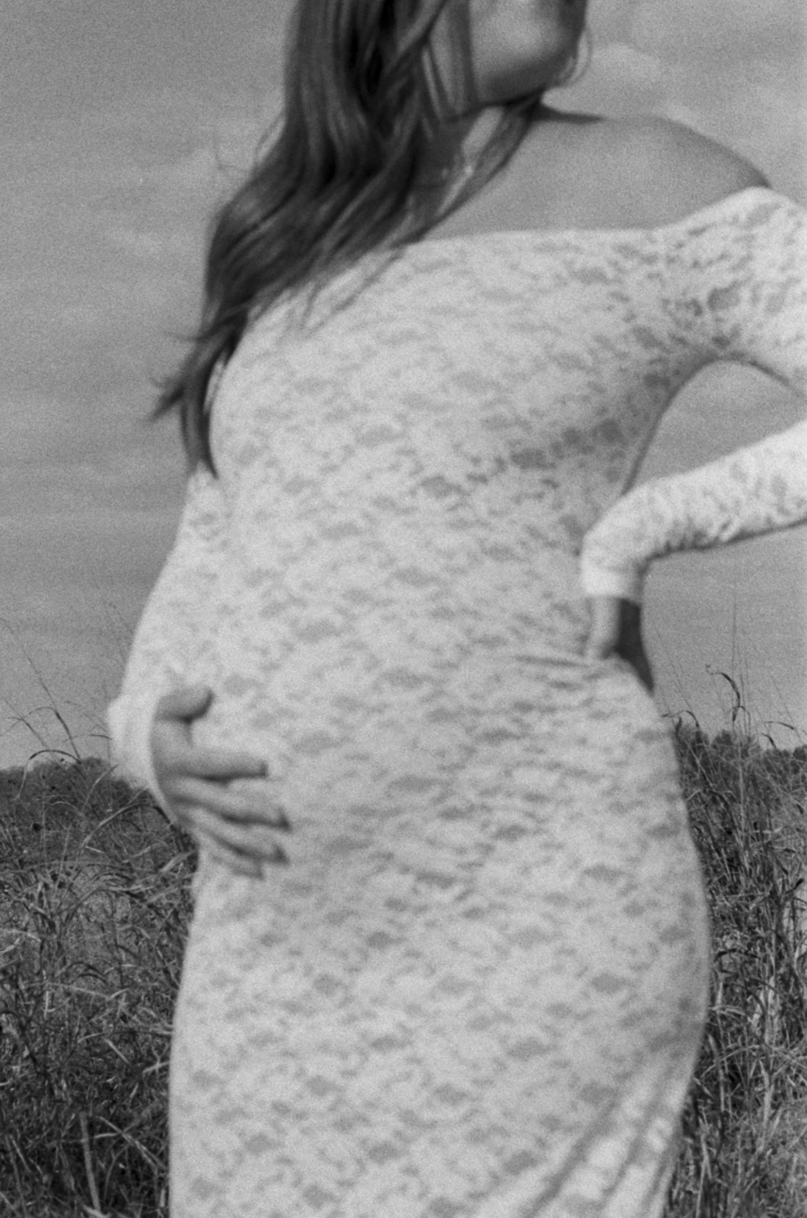 A woman in a form-fitting lace dress standing outdoors in a field, touching her pregnant belly with her right hand, her face is out of frame.