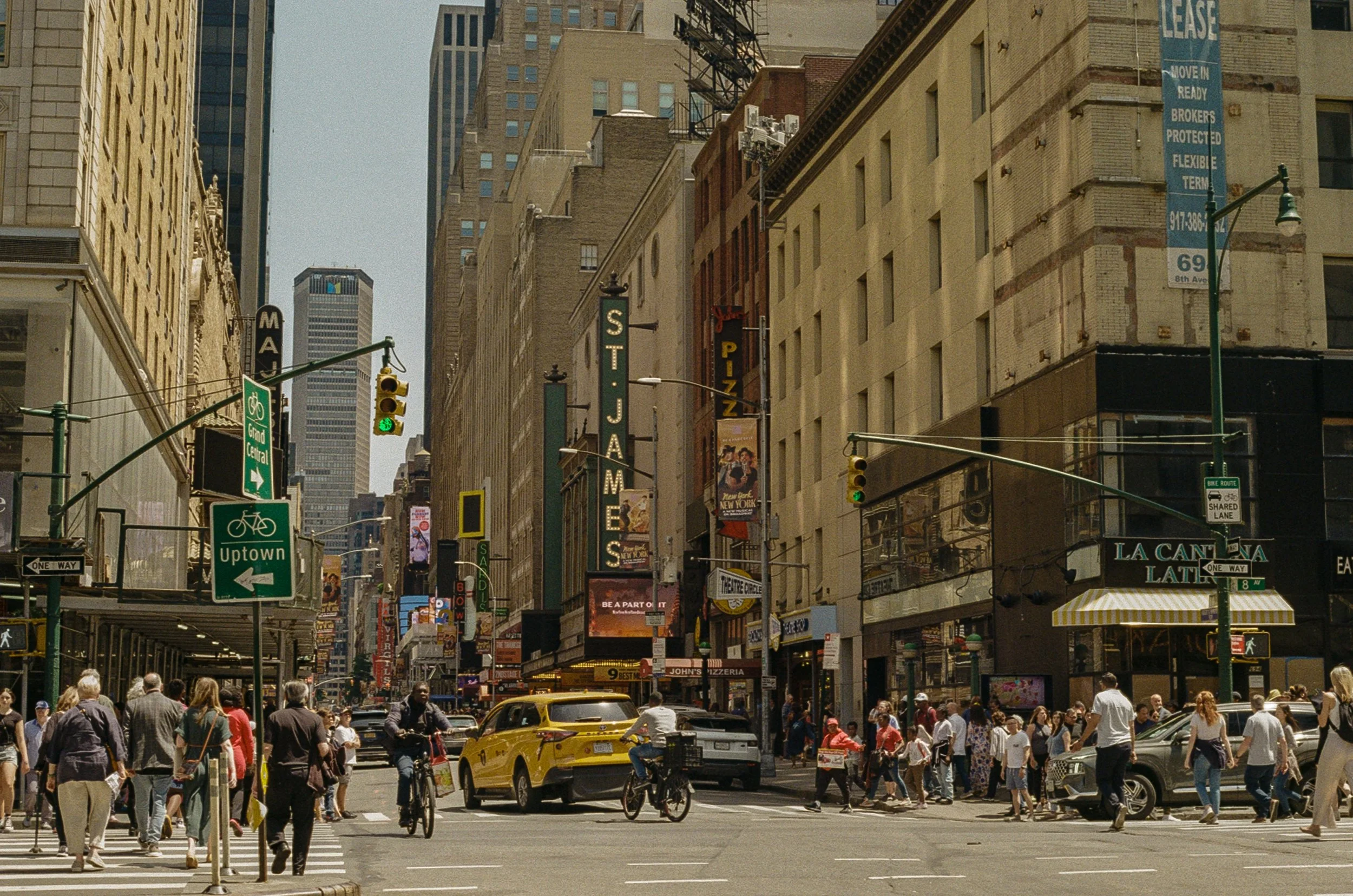 Busy city street in New York City with pedestrians crossing, a yellow taxi, shops, and tall buildings, including the St. James Theatre sign.