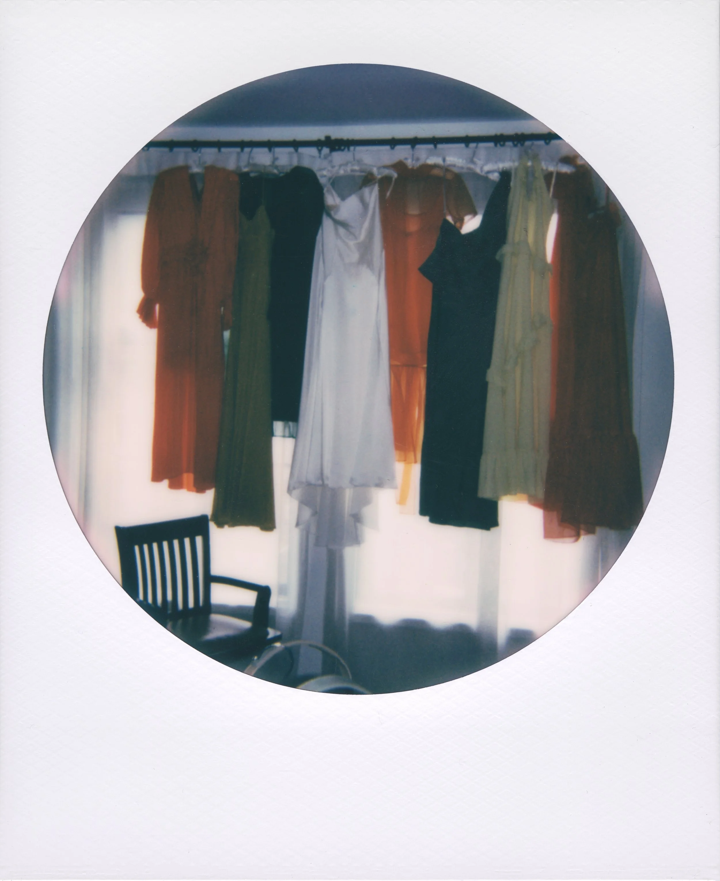 Round-frame polaroid of various dresses hanging on a clothing rack in front of a window with curtains, with a black chair partially visible in the lower left corner.