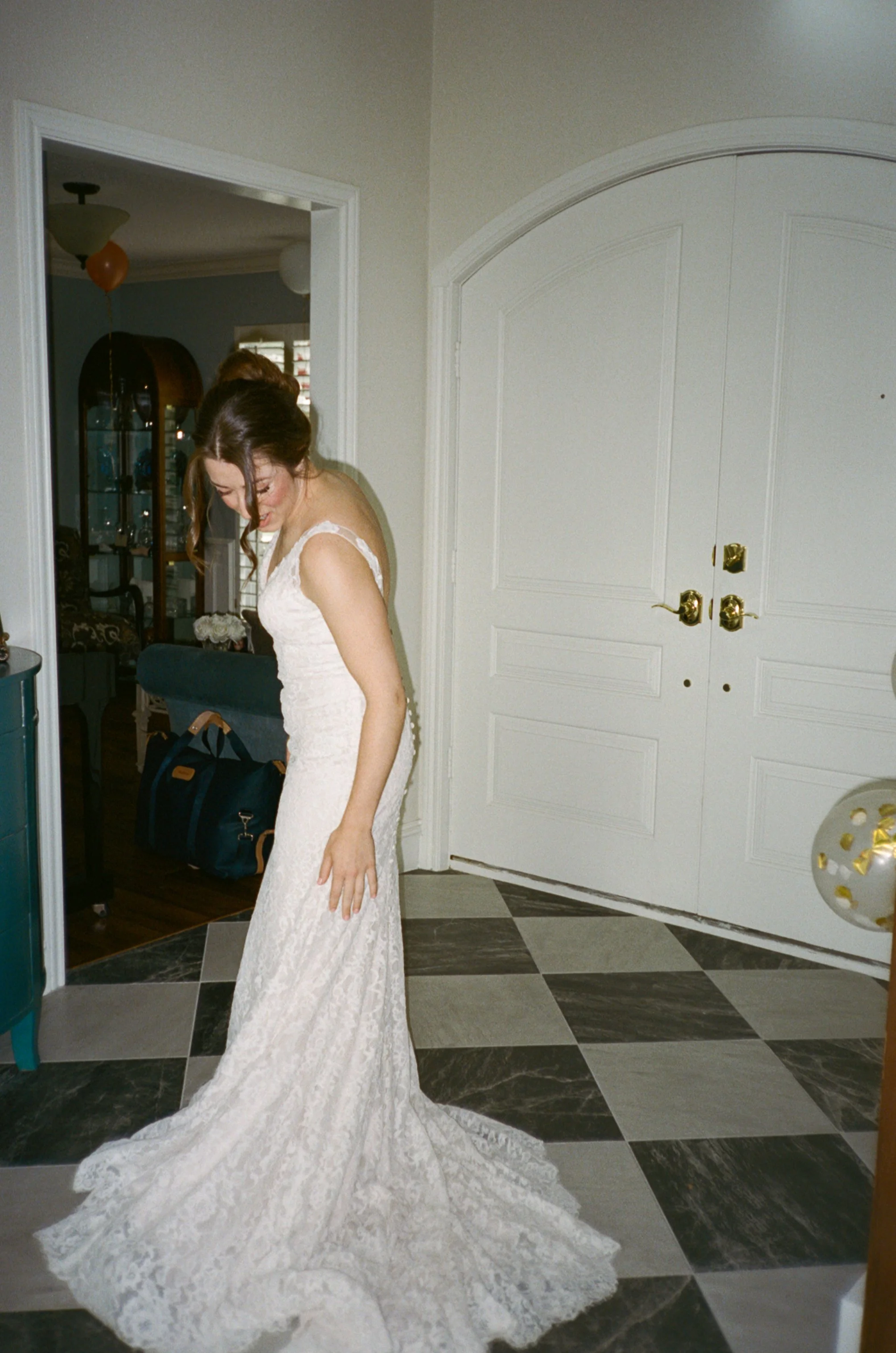 A woman in a wedding dress standing on a checkered tile floor in front of a white double door, smiling and looking down.