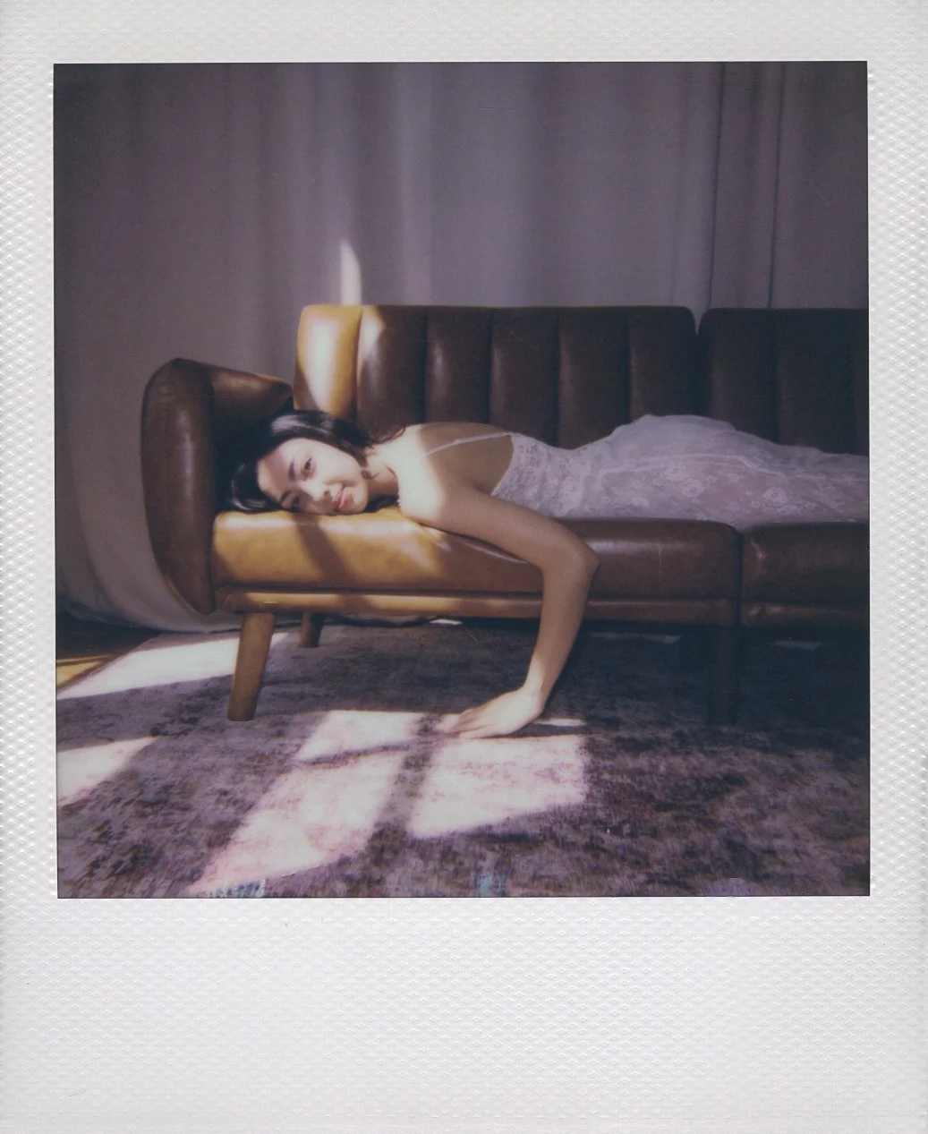Square-frame polaroid of a woman lying on a vintage brown couch in a dimly lit room with gray curtains, with sunlight creating patterns on the floor.