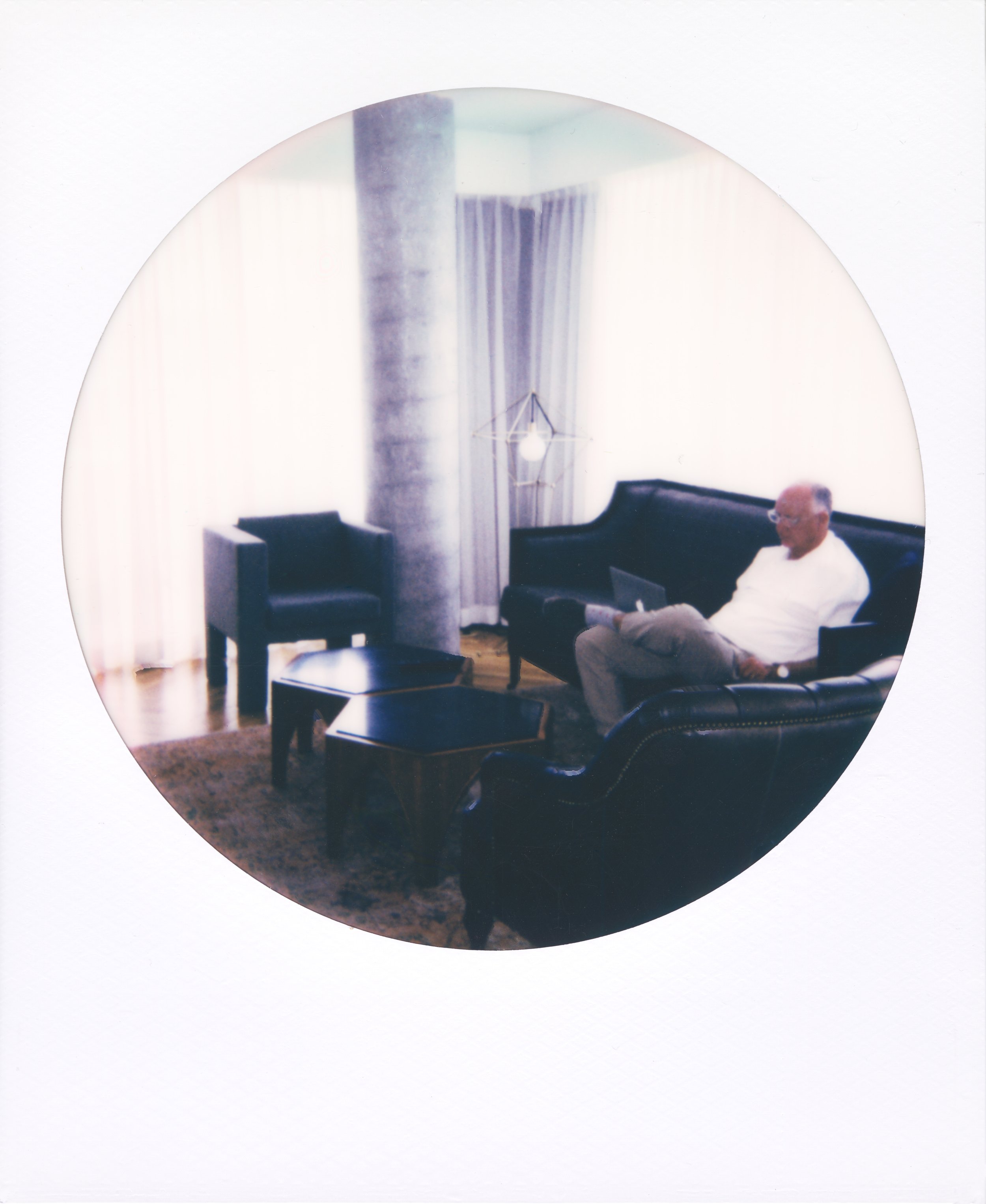 Round-frame polaroid of a man sitting on a black leather couch using a laptop, in a living room with a black armchair, a black coffee table, curtains, a spotlight, and a decorative lamp.