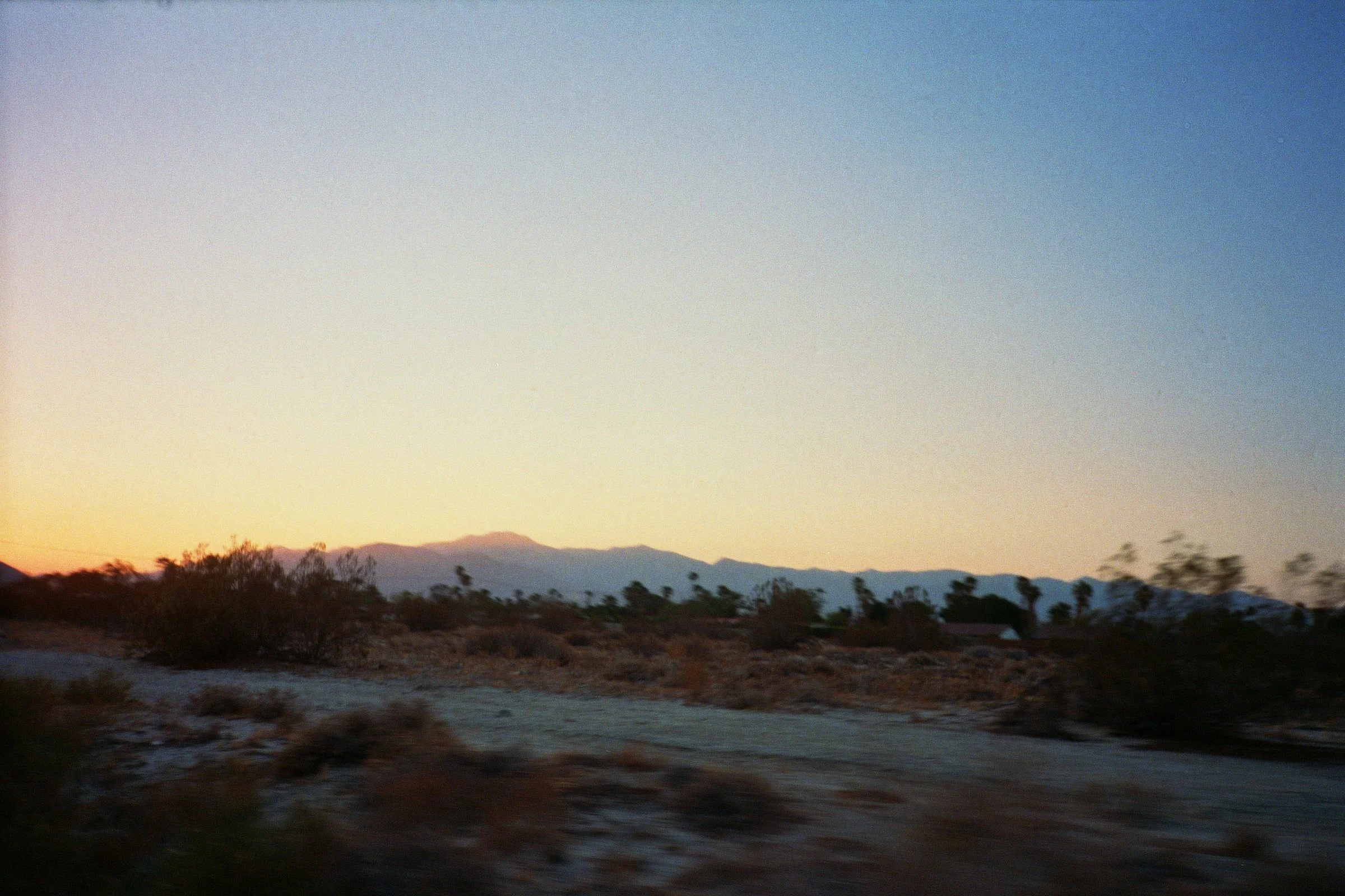 Desert landscape during sunset with mountains in the distance, sparse vegetation, and a dirt road in the foreground.