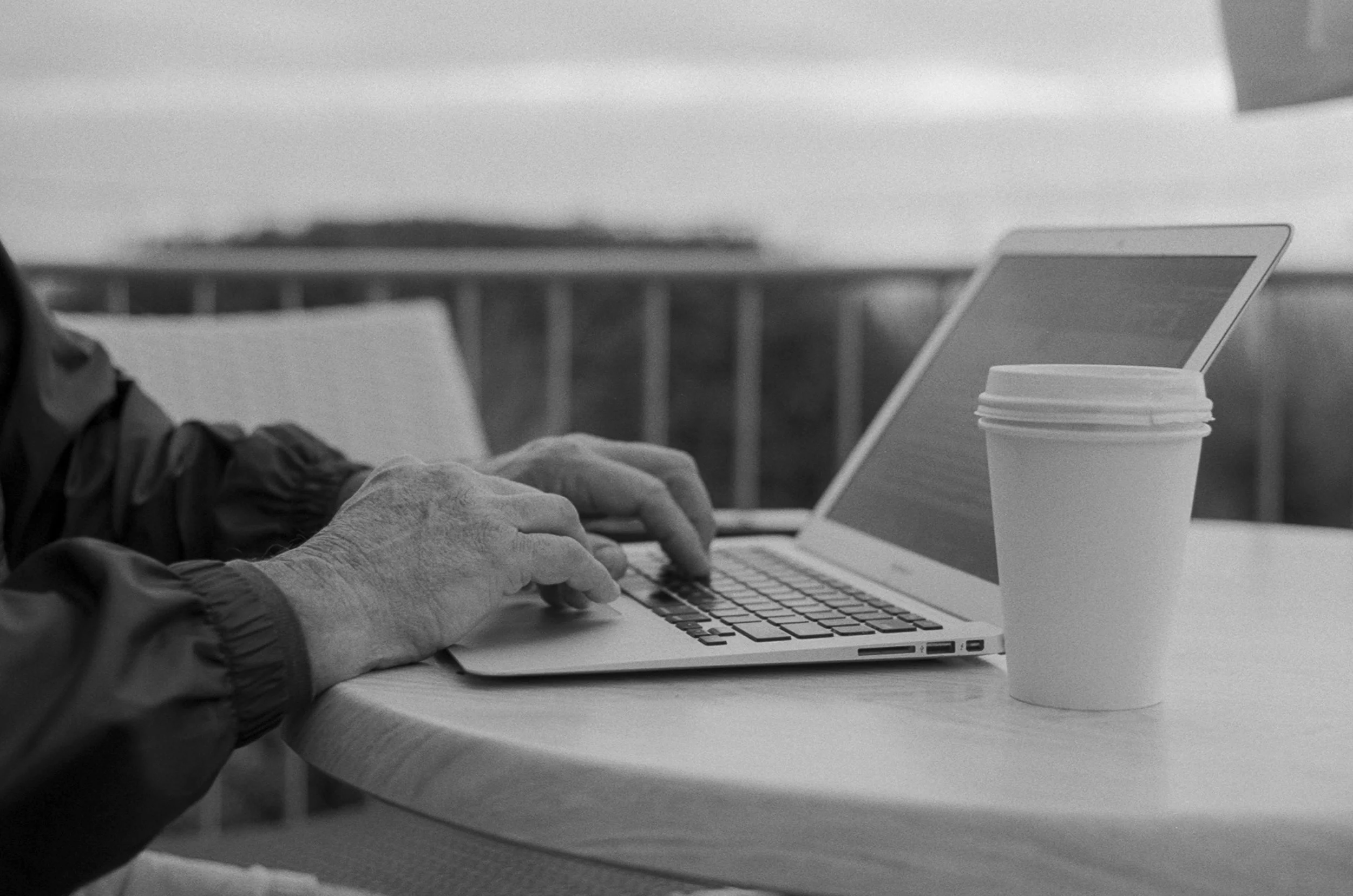 Hand of a person typing on a laptop at a table, with a takeout coffee cup nearby. 