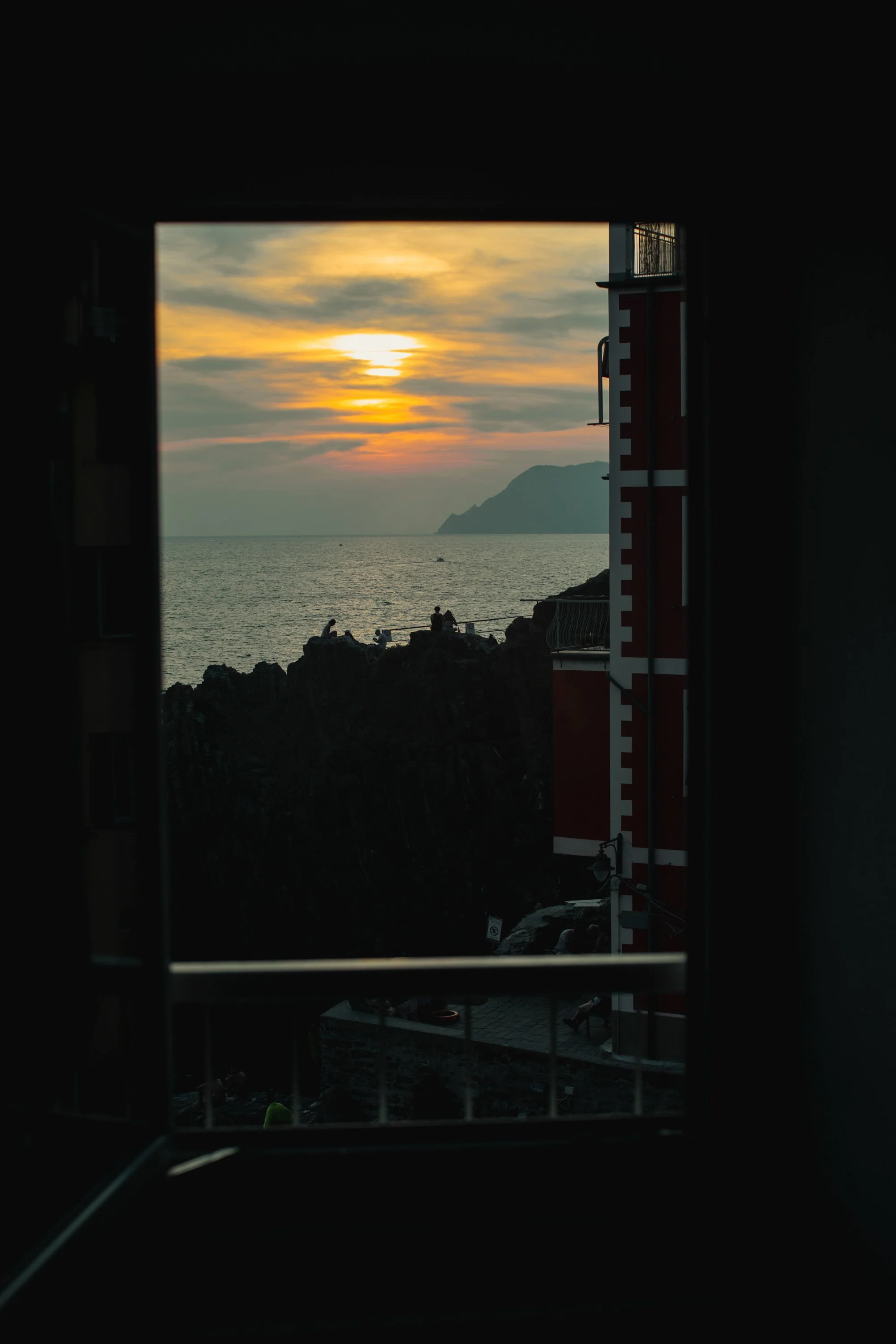Sunset over the ocean viewed through a window, with silhouetted rocks and people sitting on the rocks, a distant landmass, and a partly cloudy sky.