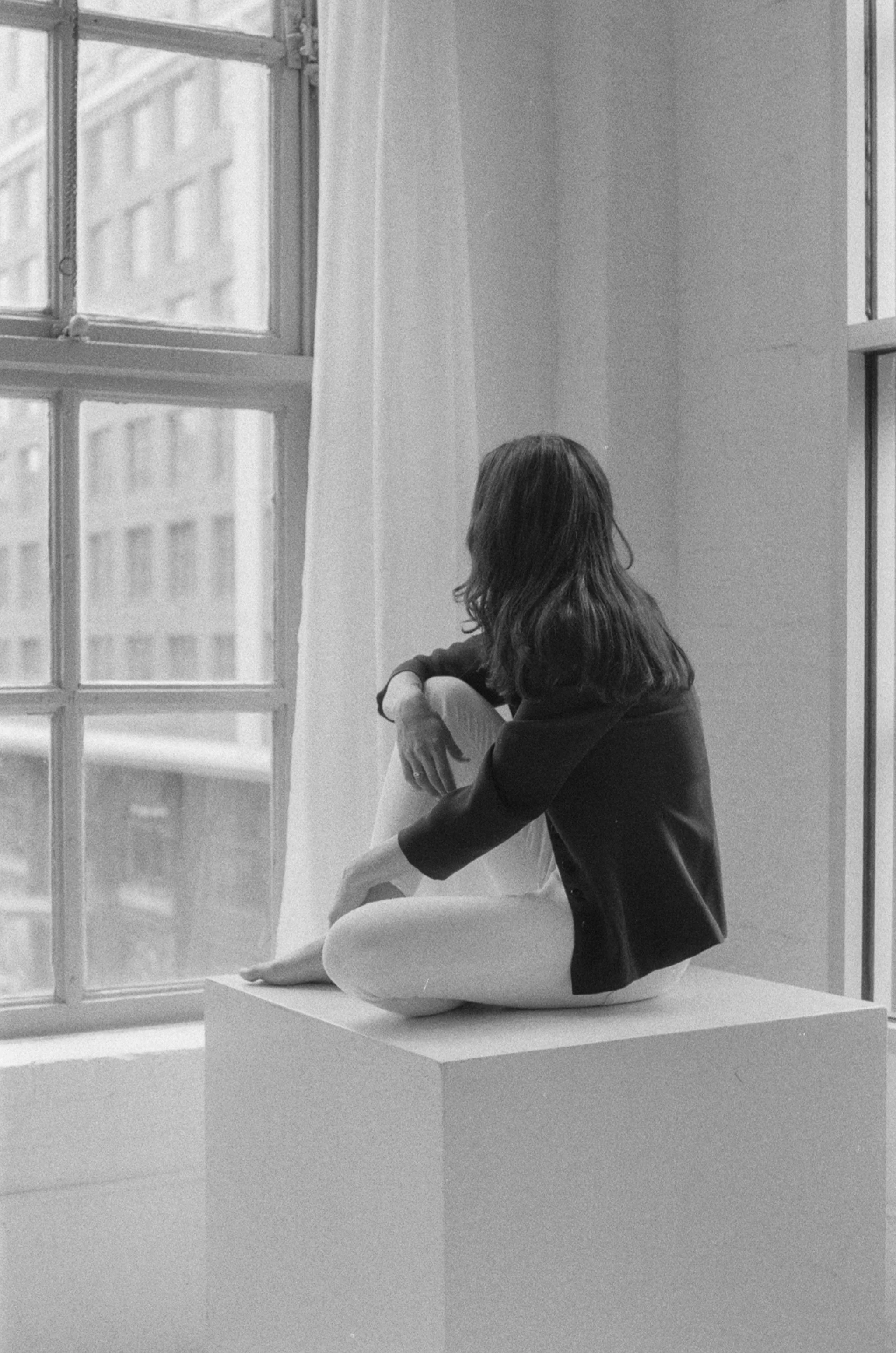 A woman sitting on a podium near large windows, looking outside in an indoor setting.