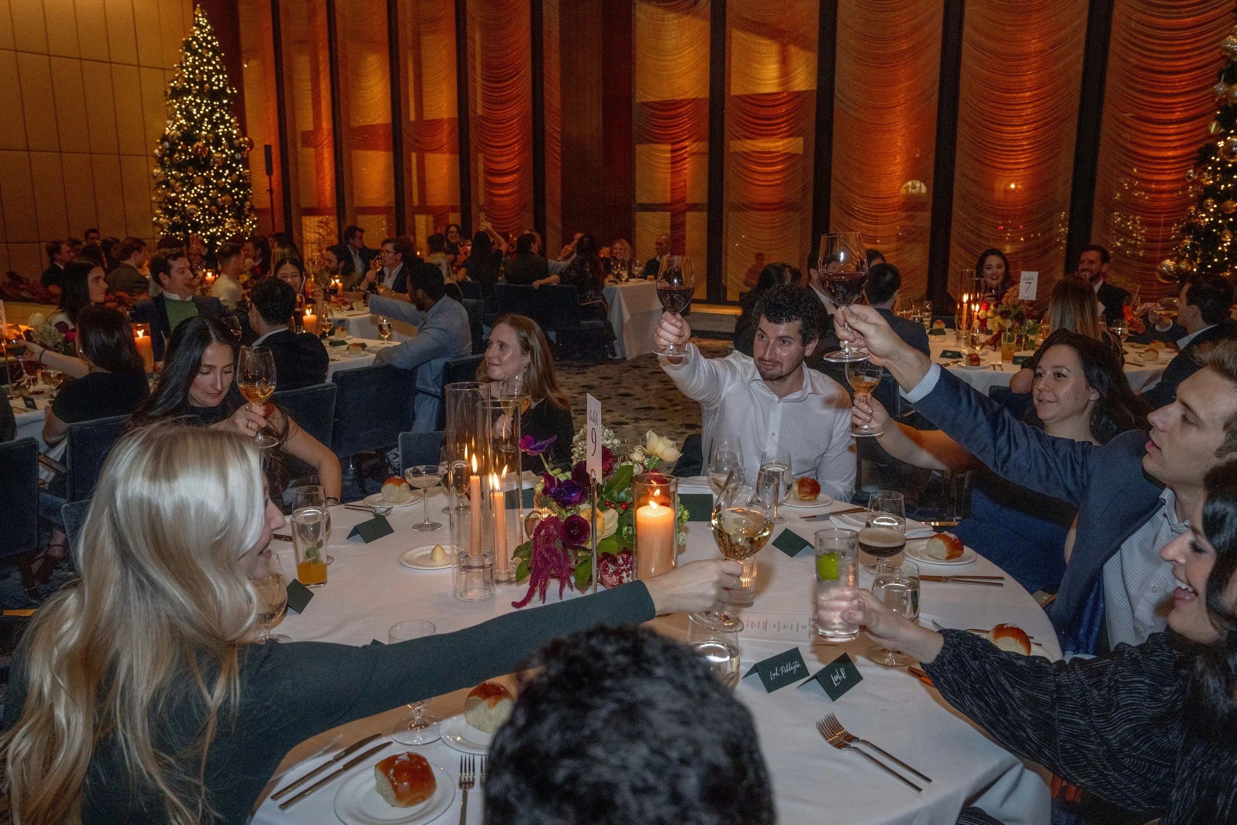 People dressed in formal attire celebrating at a Christmas dinner in a decorated banquet hall with Christmas trees and warm lighting, raising glasses for a toast.