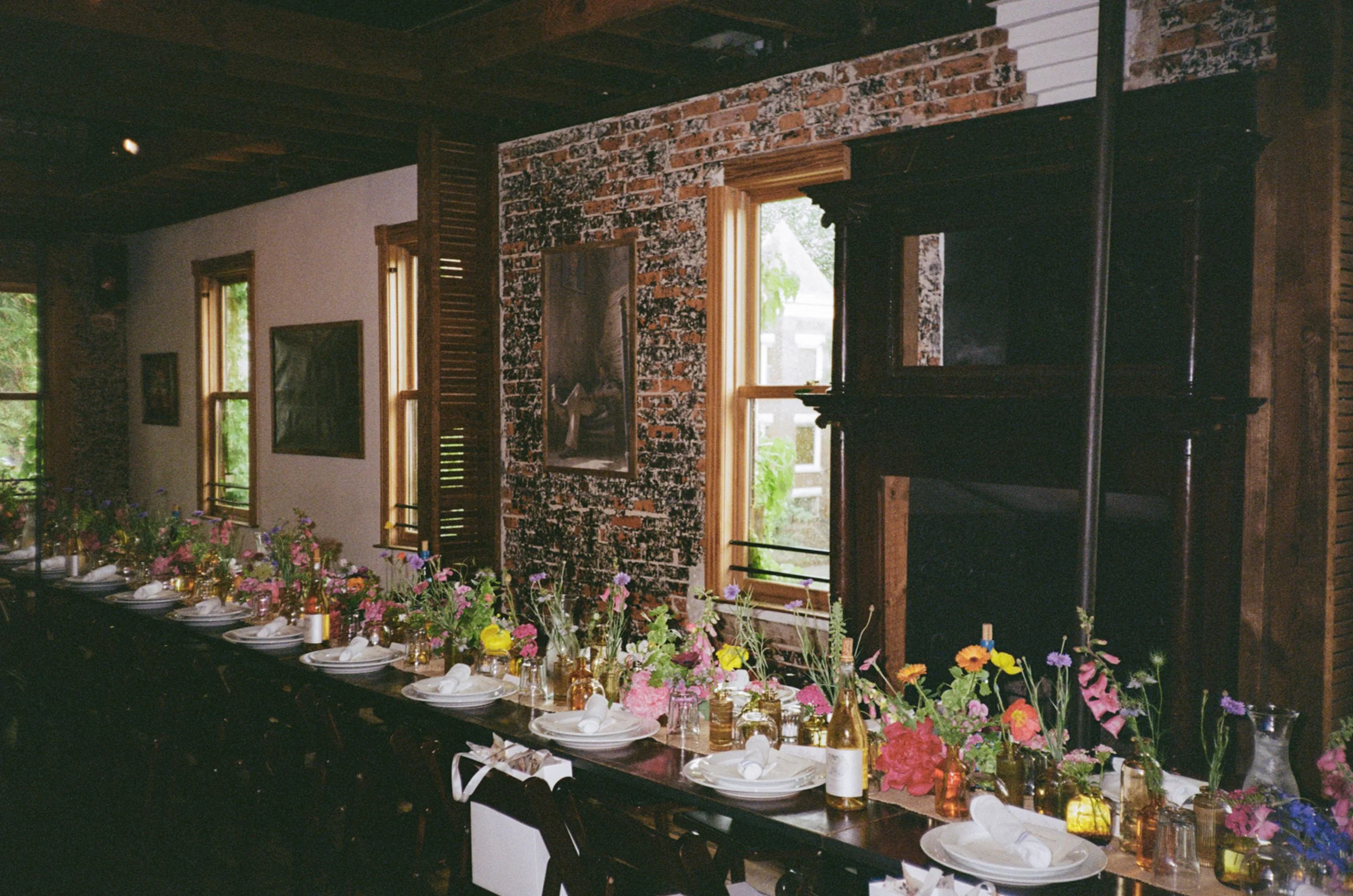 Long dining table decorated with colorful flowers in vases, set with white plates, silverware, and napkins, in a rustic room with brick wall and wooden framed windows.