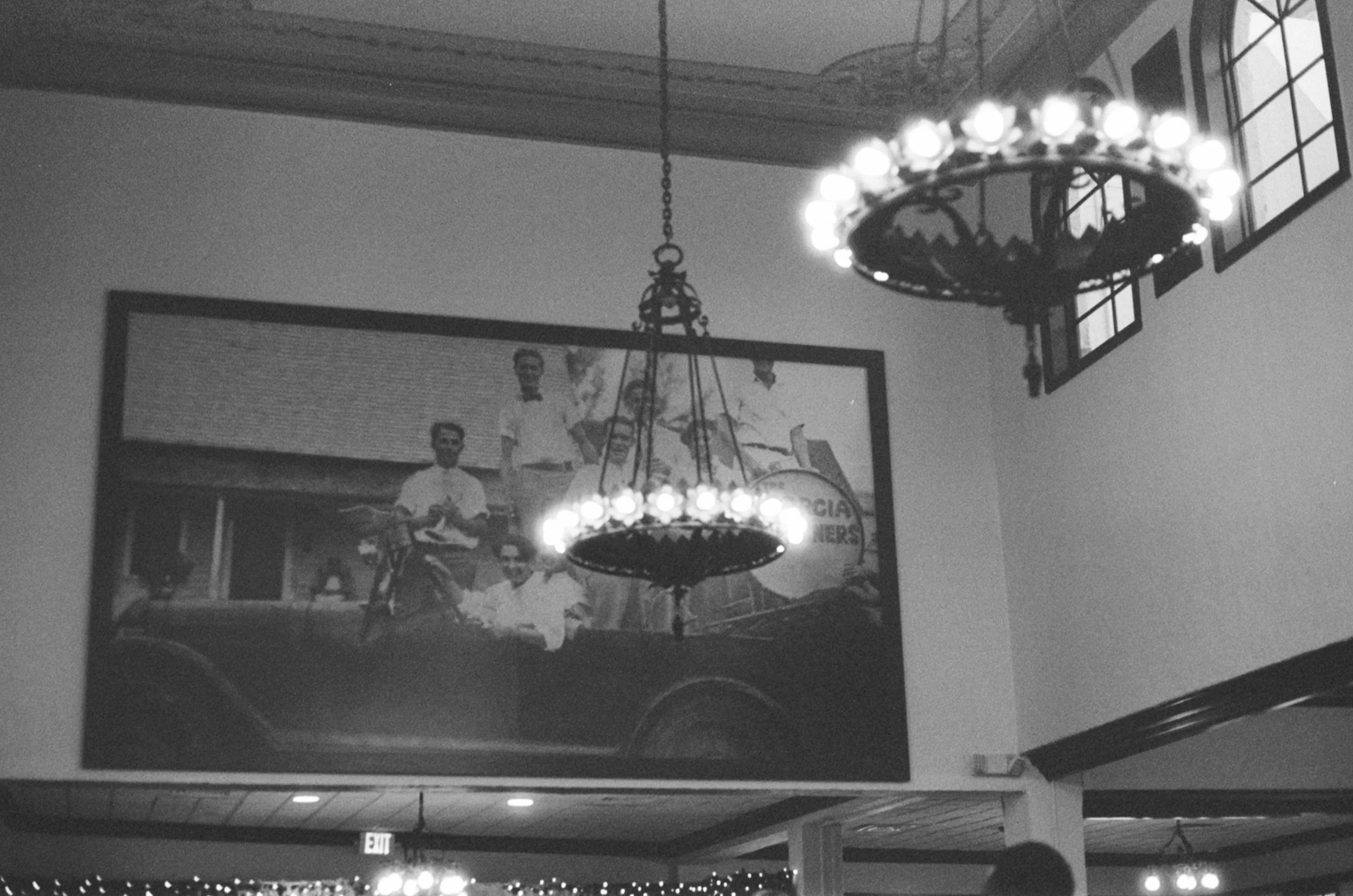 Black and white photo of a dining room with a large portrait of five men on the wall, two chandeliers hanging from the ceiling, and a row of windows along the top edge.