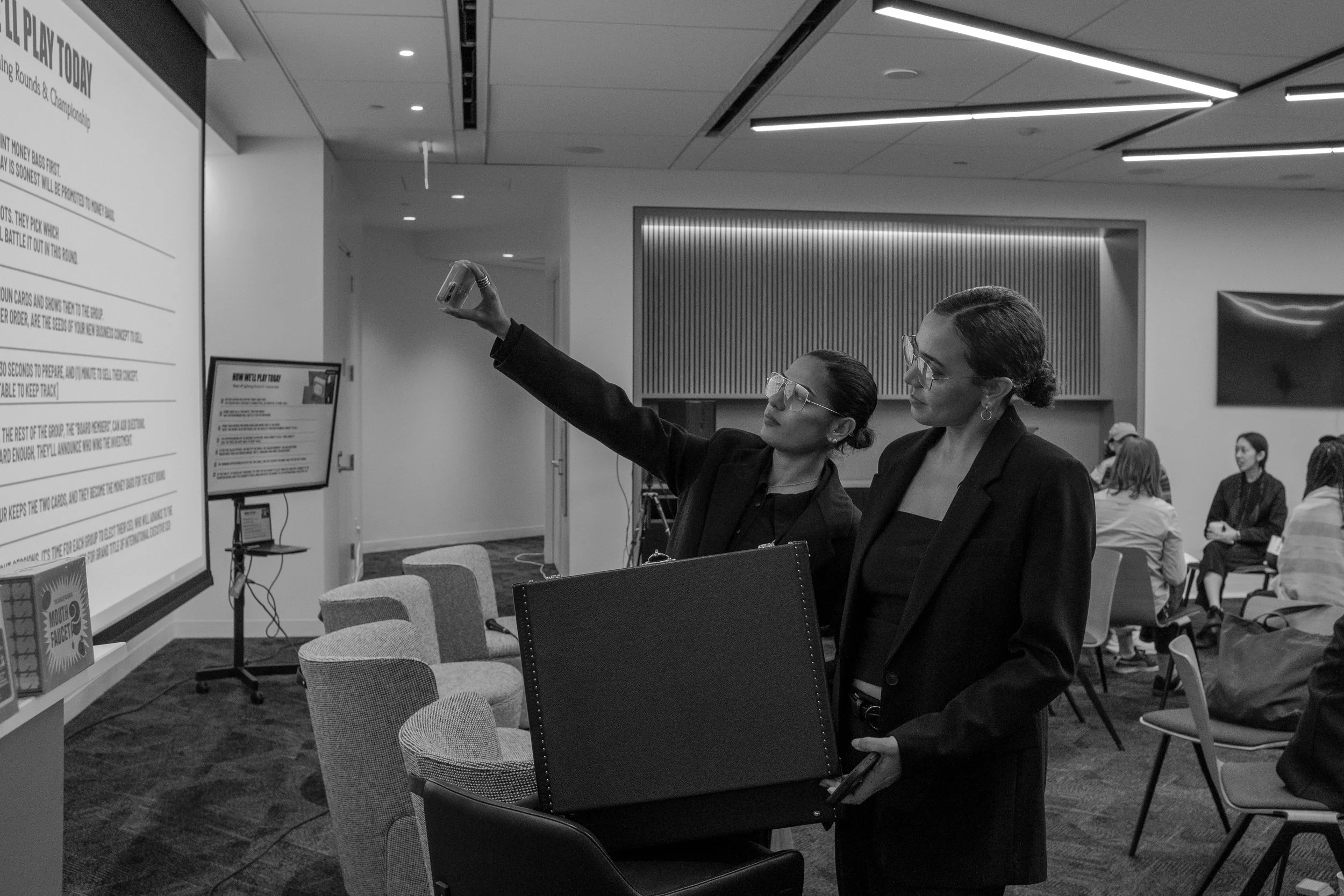 Two women in business attire, wearing glasses, are taking a selfie in a conference room. Several people are seated in the background.