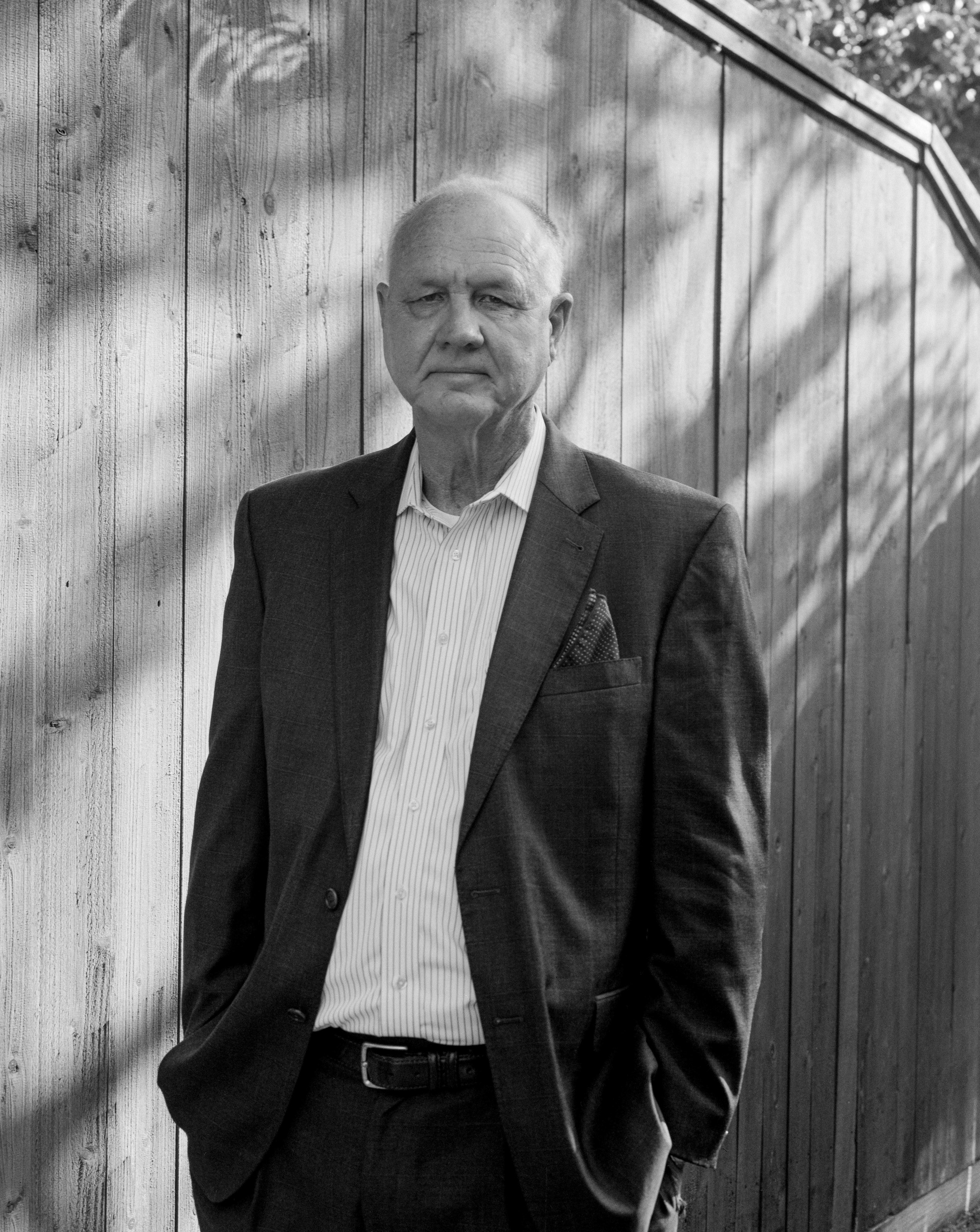 A black and white photo of a man in a suit standing outdoors against a wooden fence.