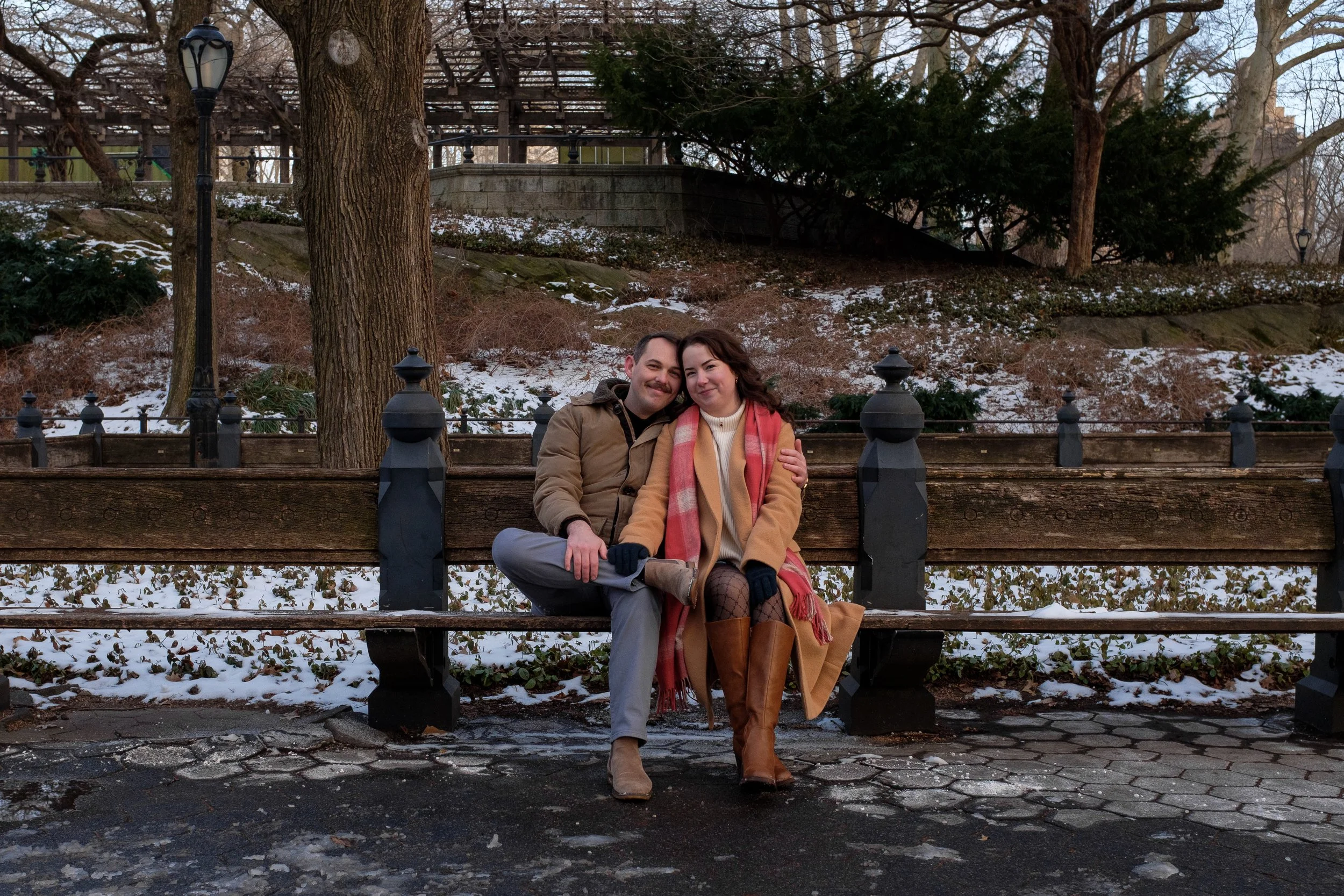 A couple sitting on a park bench during winter, smiling and cuddling with snow on the ground and trees in the background.