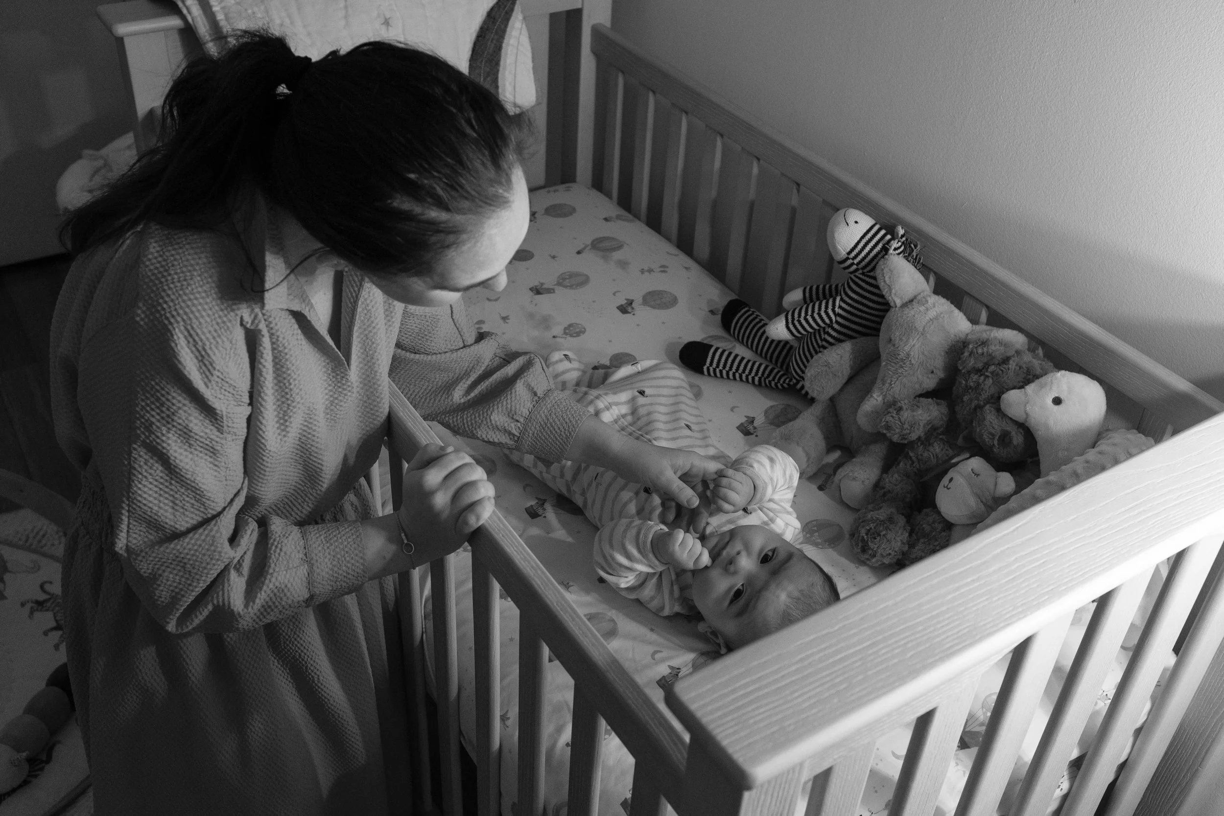 A woman leaning over a crib, looking at a baby lying on its back surrounded by stuffed animals.