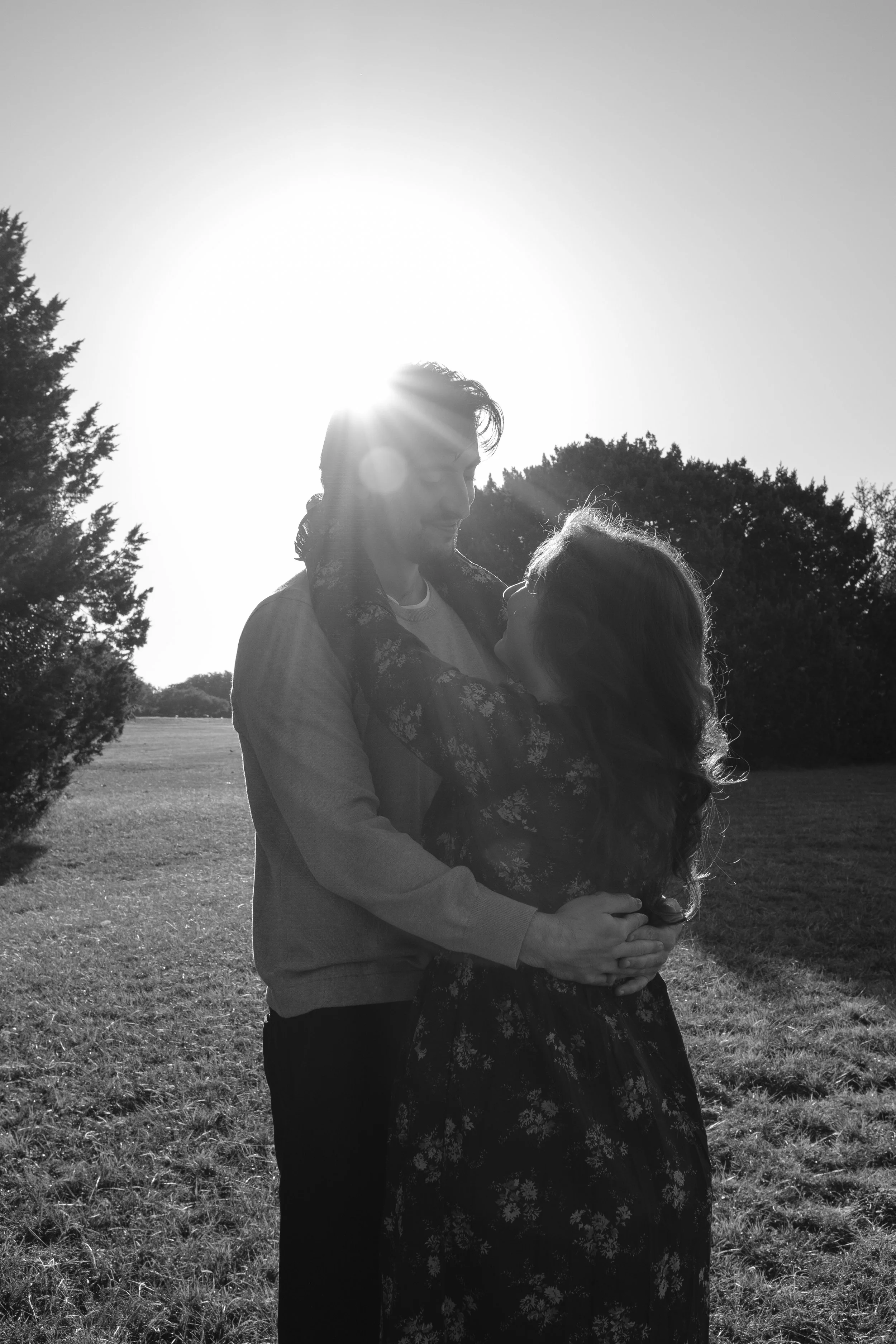A black and white photo of a couple standing outdoors in a field, embracing each other with the sun setting behind them.
