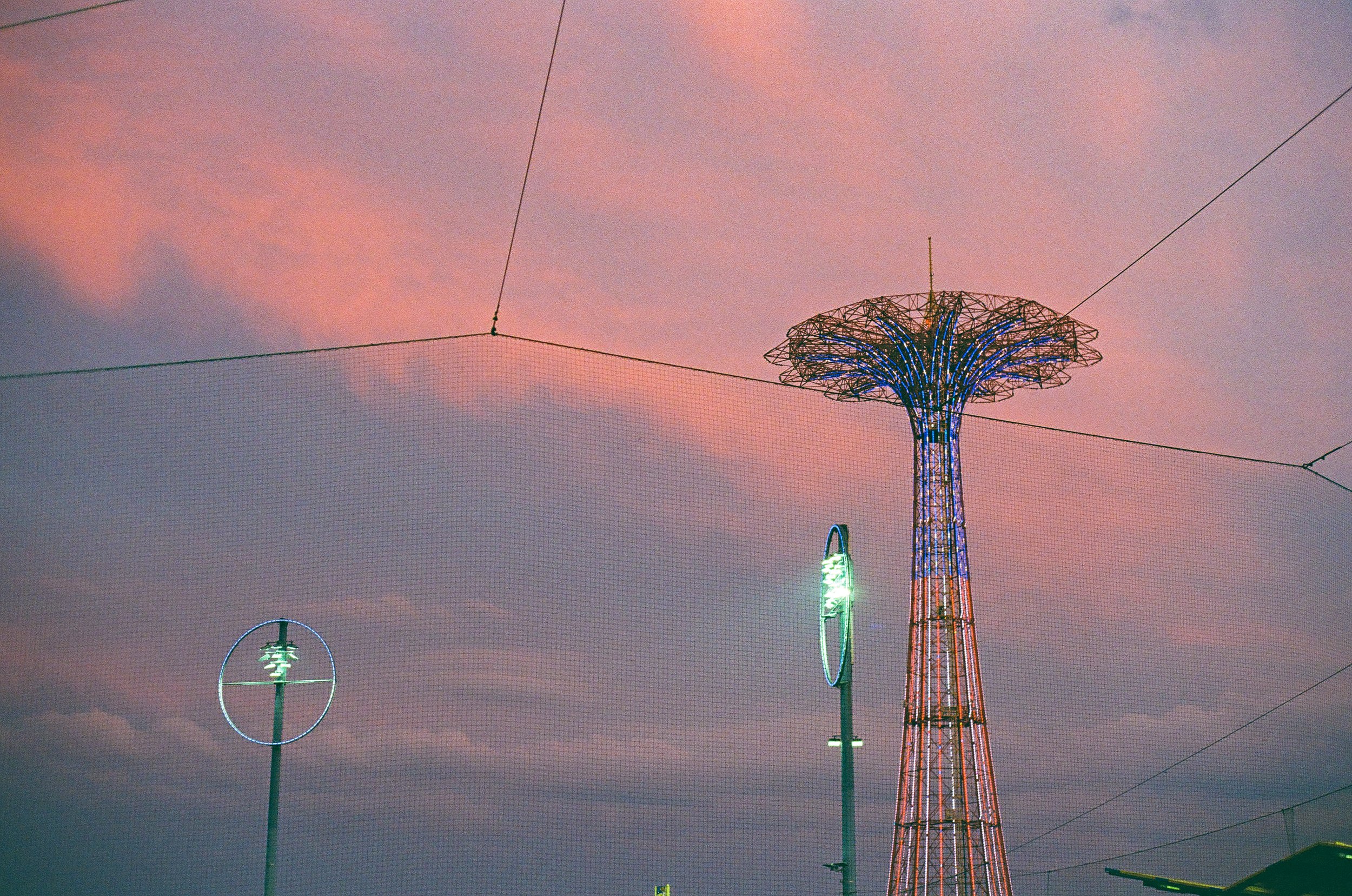 A tall tower with a decorative top lit with blue lights, set against a pink and purple sunset sky. There are also streetlights and wires visible in the foreground.