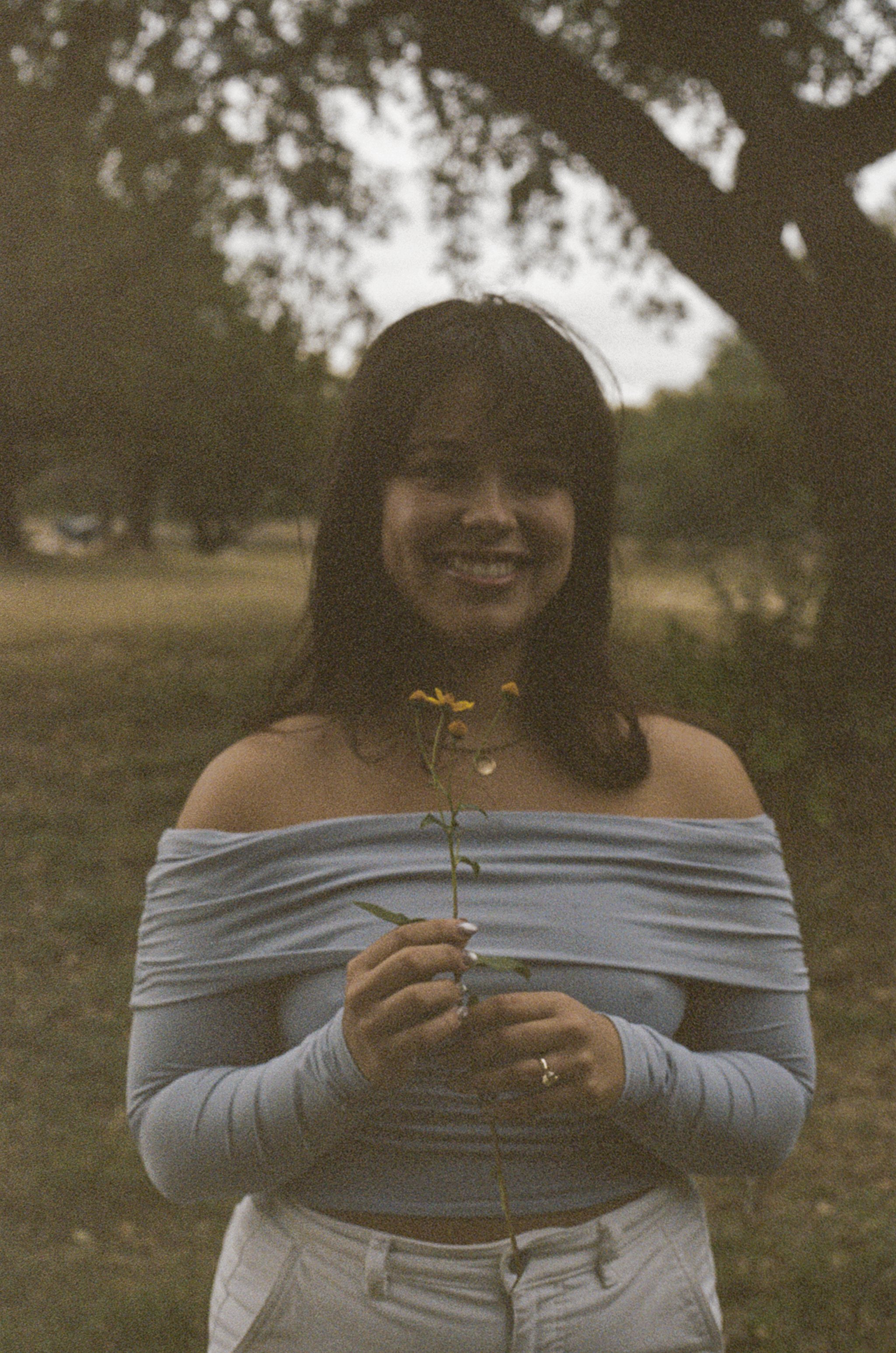 A woman standing outdoors under a tree, smiling, holding a small yellow flower, wearing an off-shoulder blue top and white pants.