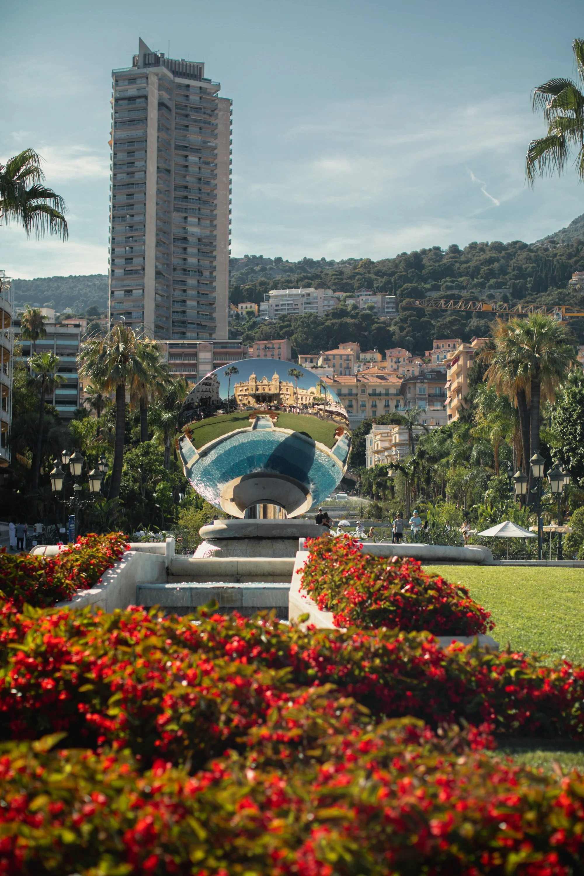 City park with modern buildings, palm trees, colorful flowers, and a reflective metallic sculpture in the center.