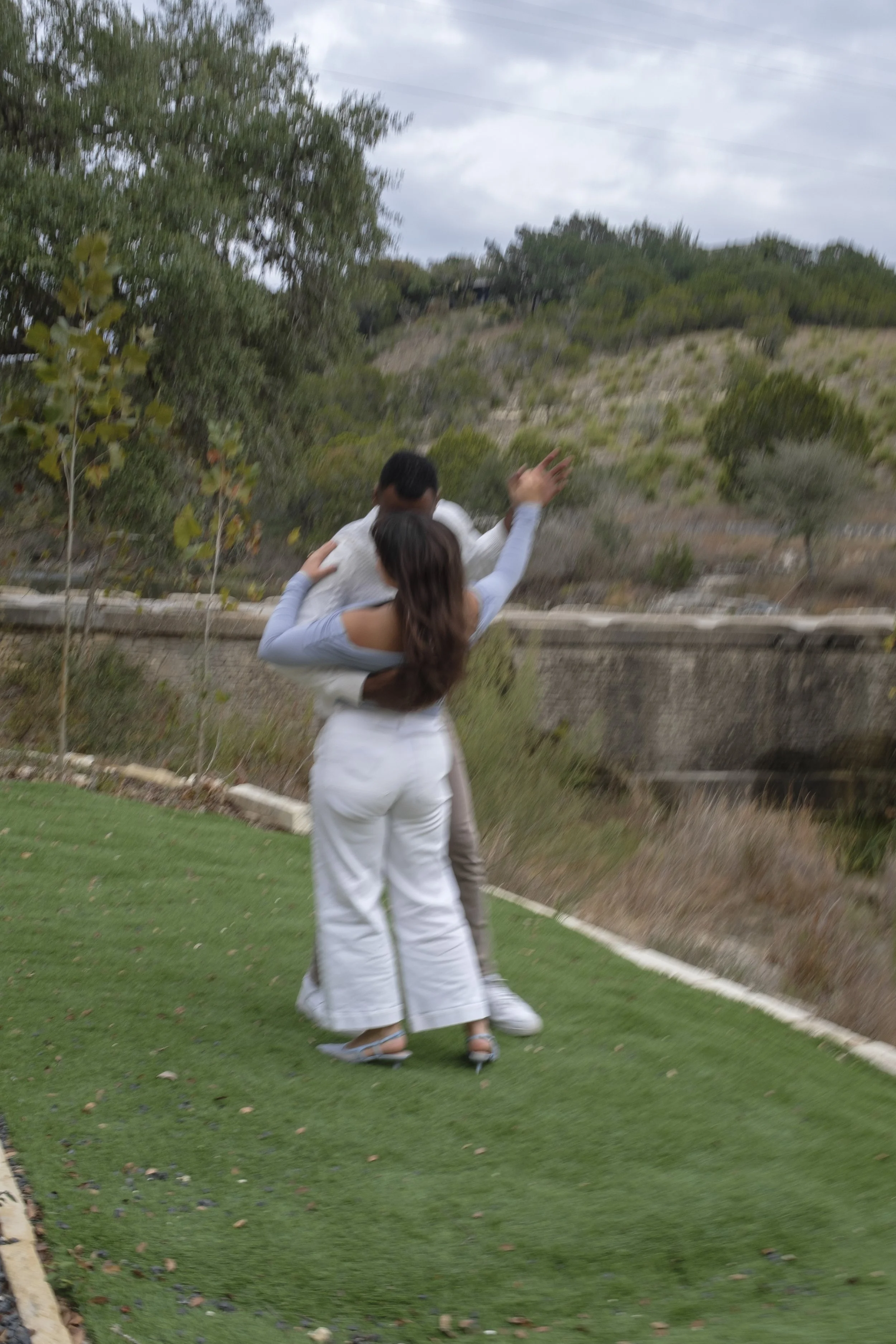 A woman and a man dancing outdoors on a grassy area near a pond with trees and hills in the background.