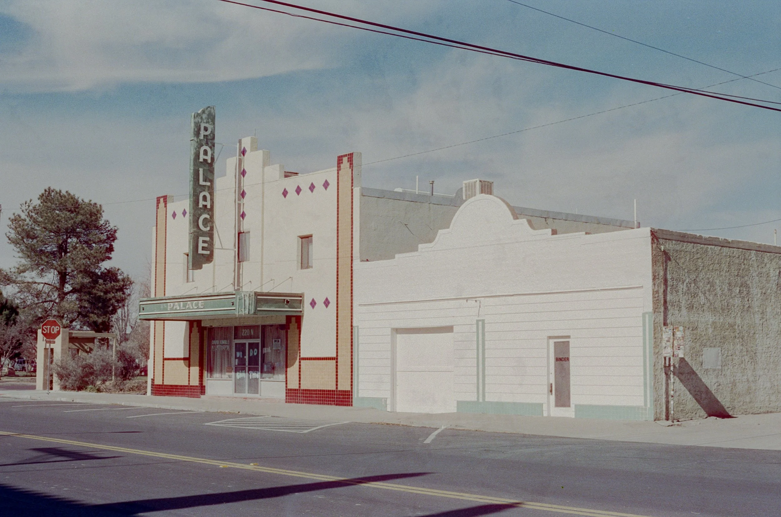 An old theater building with a vertical sign reading 'PALACE' in large letters. The building has a vintage marquee and a facade with red and white trim. There is a tree on the left and a stop sign at the corner. The sky is partly cloudy.