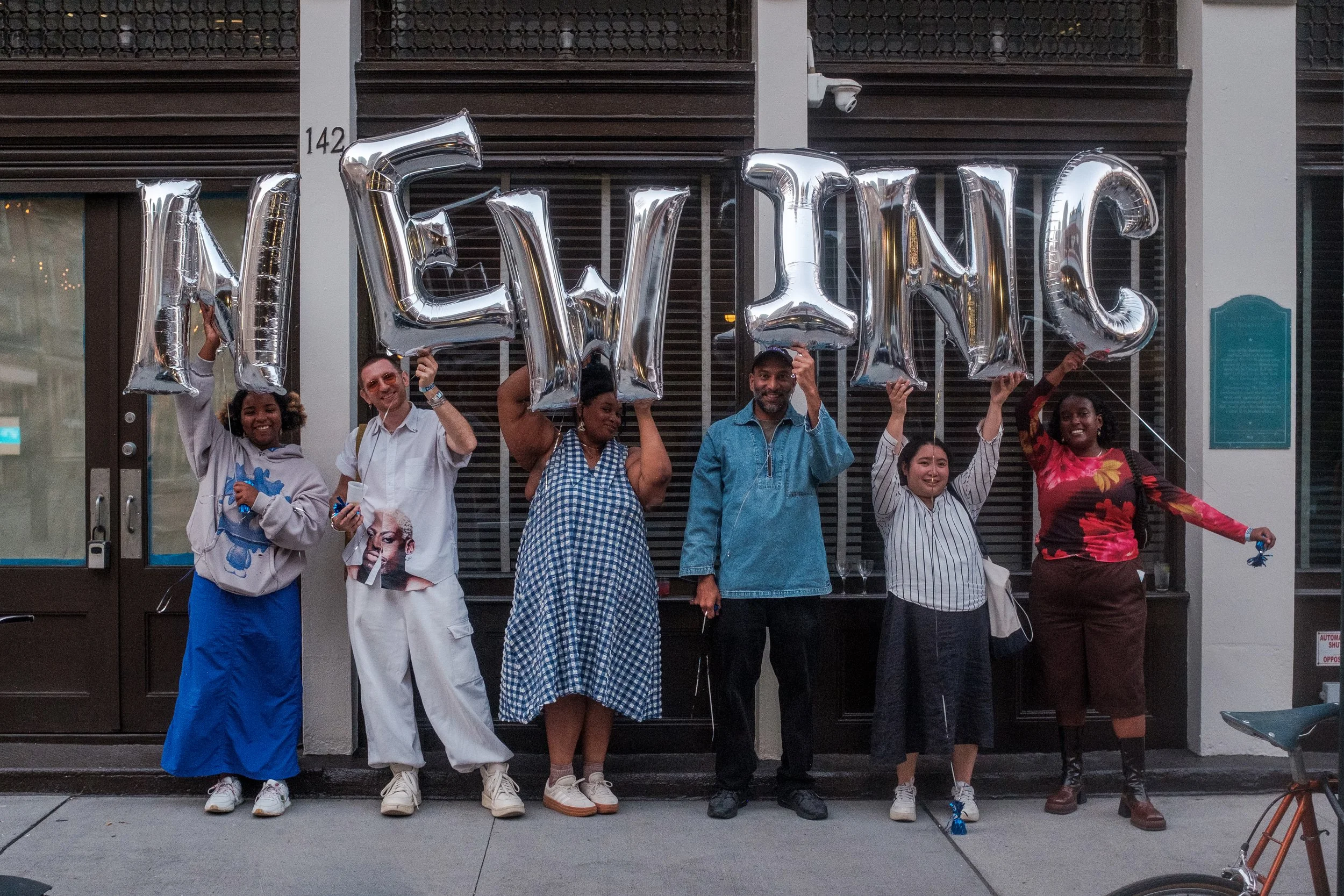 Group of six diverse people holding large silver balloons spelling 'NEW INC' outside a building.