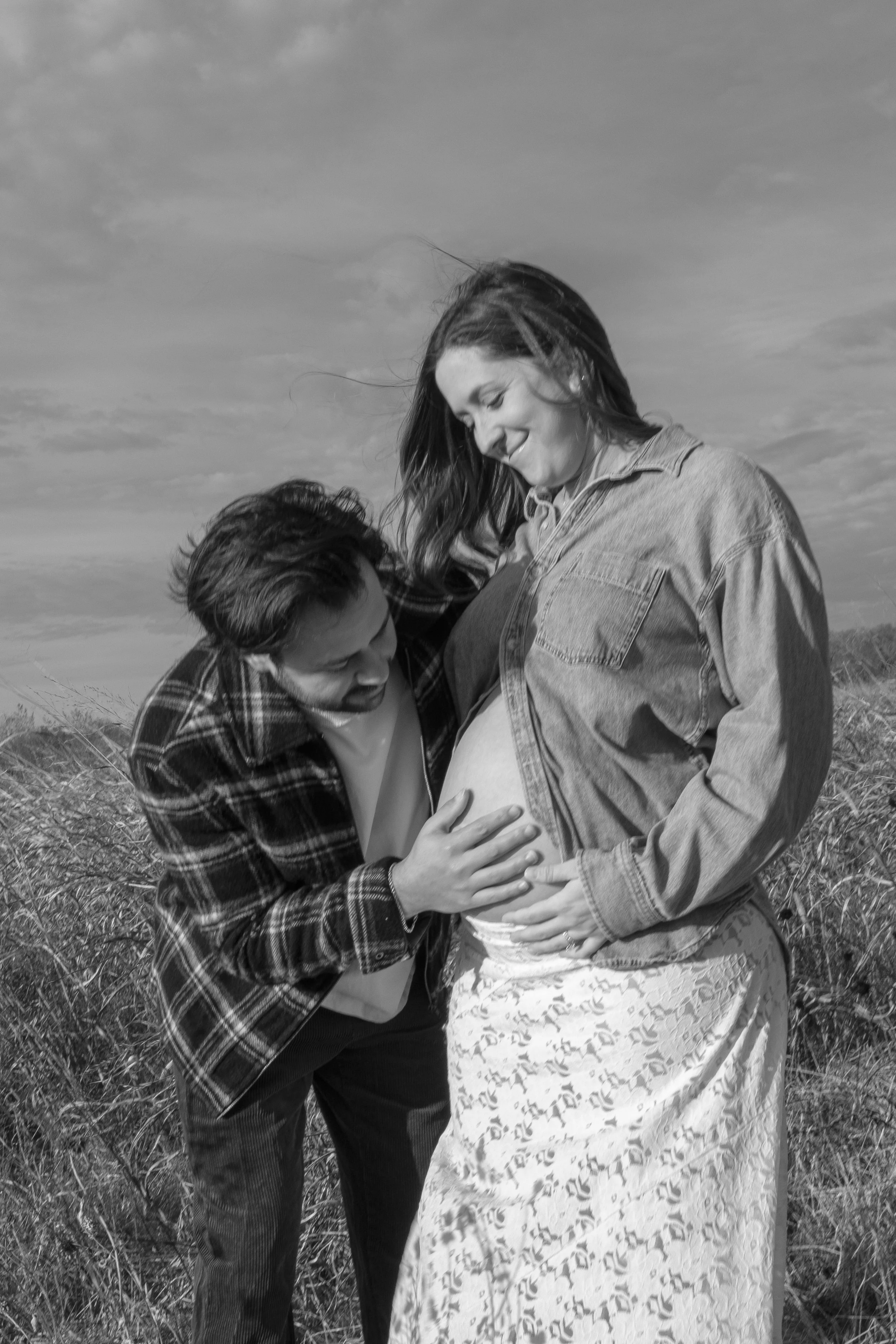 A pregnant woman standing in a field, smiling while a man touches her belly. They are outdoors with a cloudy sky overhead.