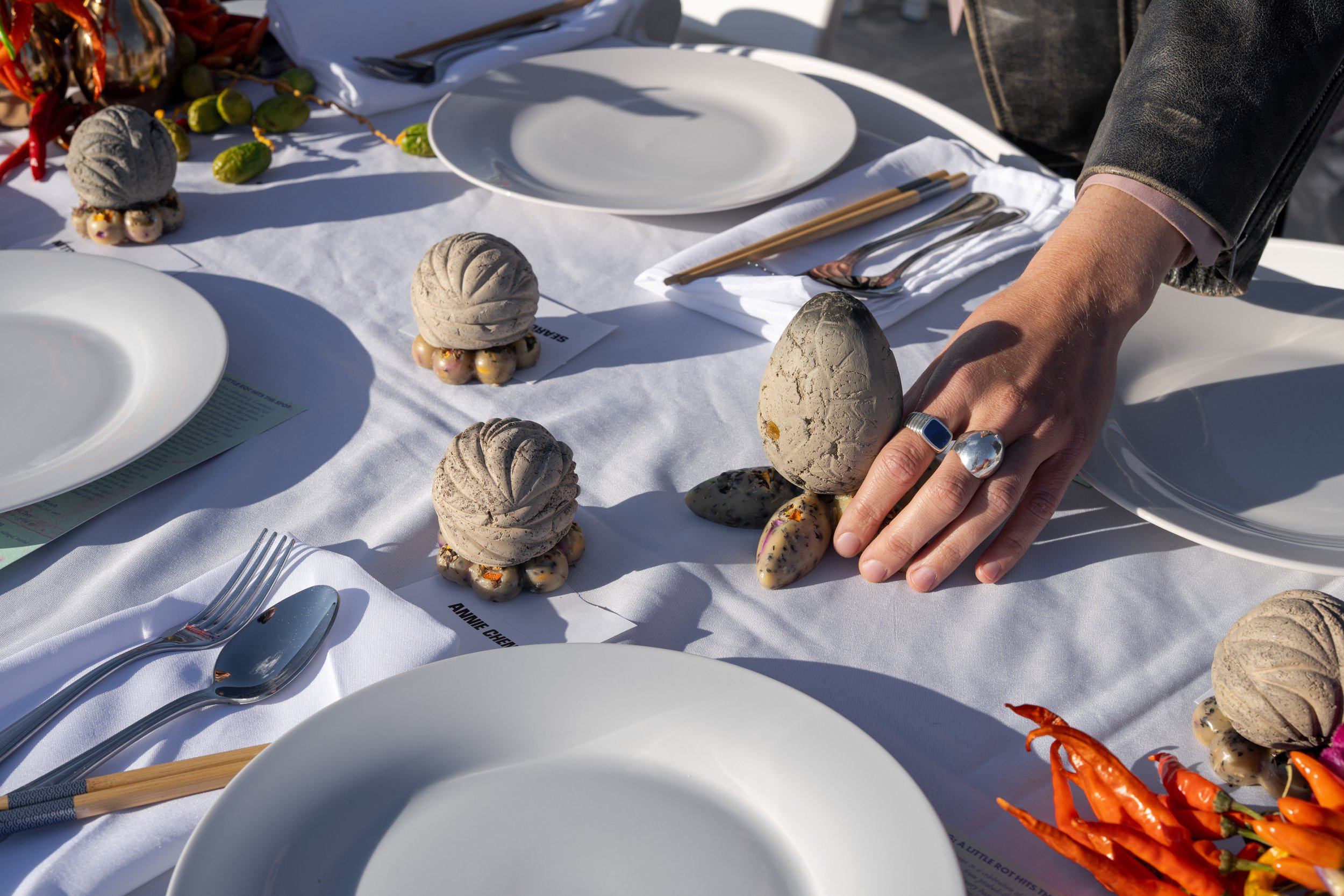 A table set with white plates, silverware, and decorative stone sculptures topped with moss, arranged on a white tablecloth during daylight. A person's hand with rings touches one of the sculptures. There are also orange chili peppers on the table.