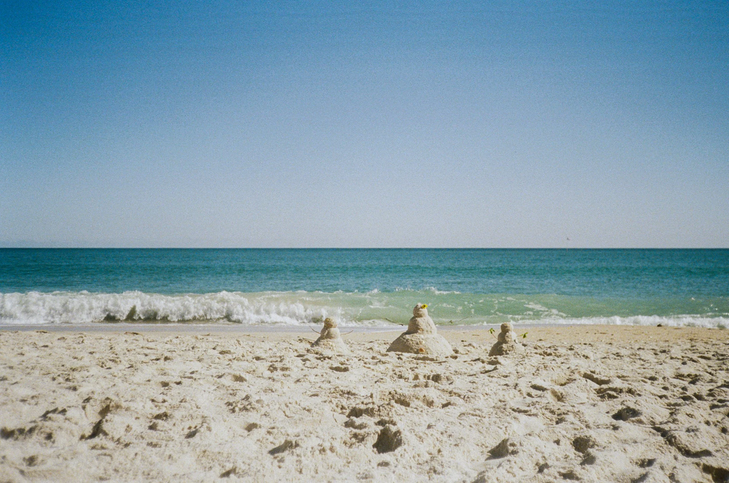 Three snowmen sandcastles on a sandy beach with ocean waves in the background and a clear blue sky.