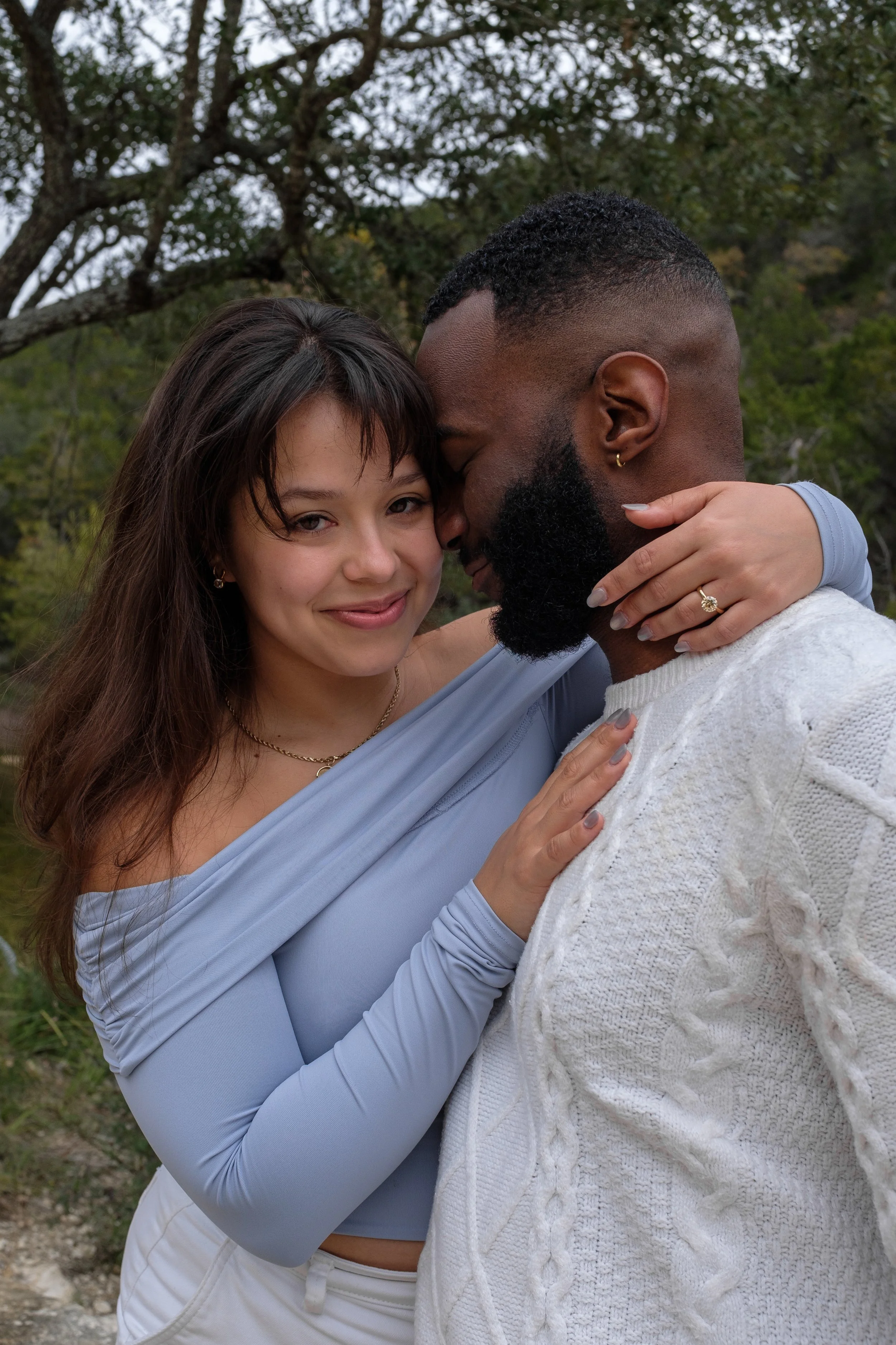 A couple embracing outdoors, with the woman smiling and the man with his eyes closed facing her, surrounded by trees.