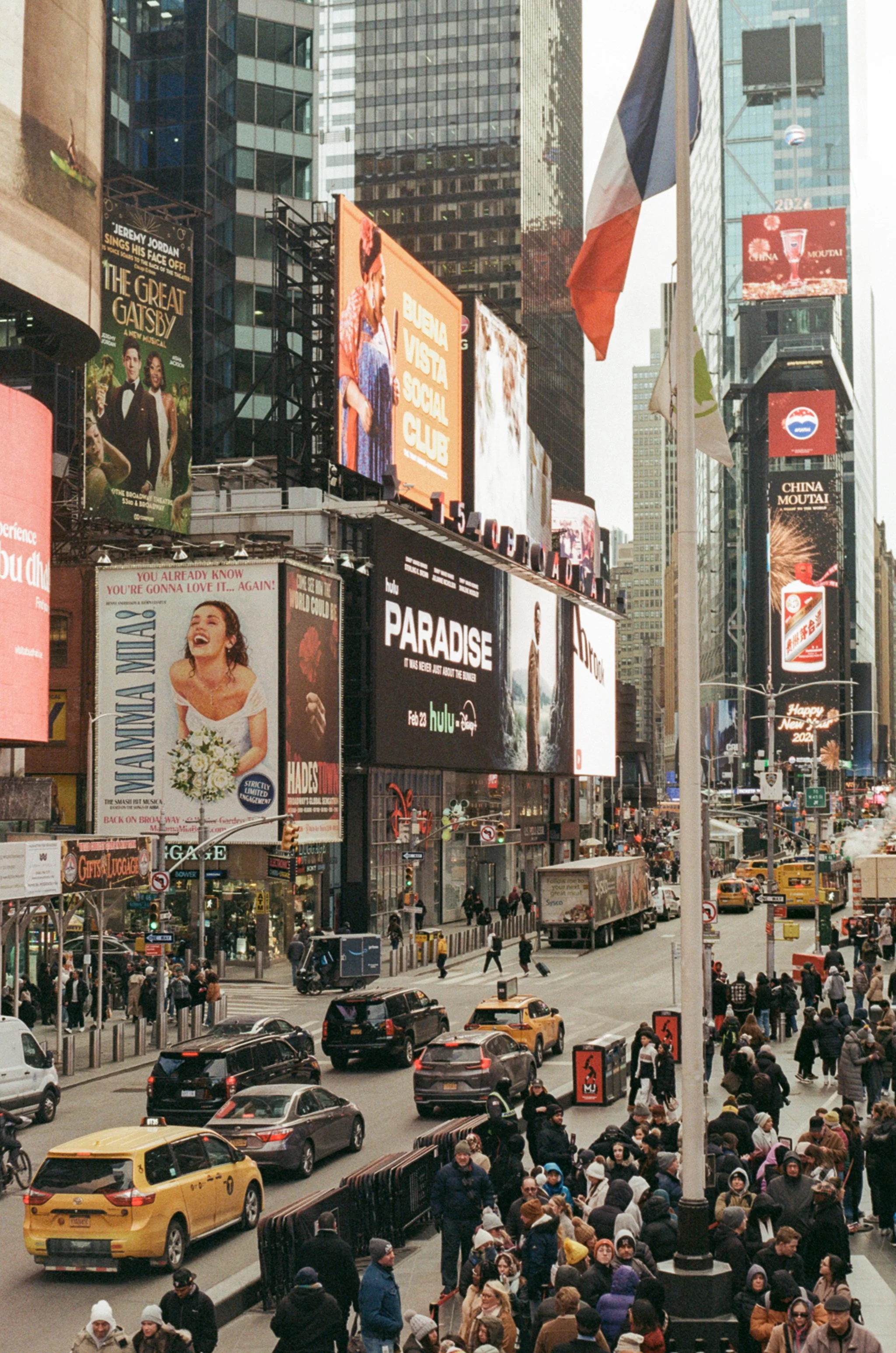Busy city street in Times Square, New York City, with crowds of pedestrians on the sidewalk, yellow taxis and cars on the road, and large digital billboards displaying advertisements and messages.