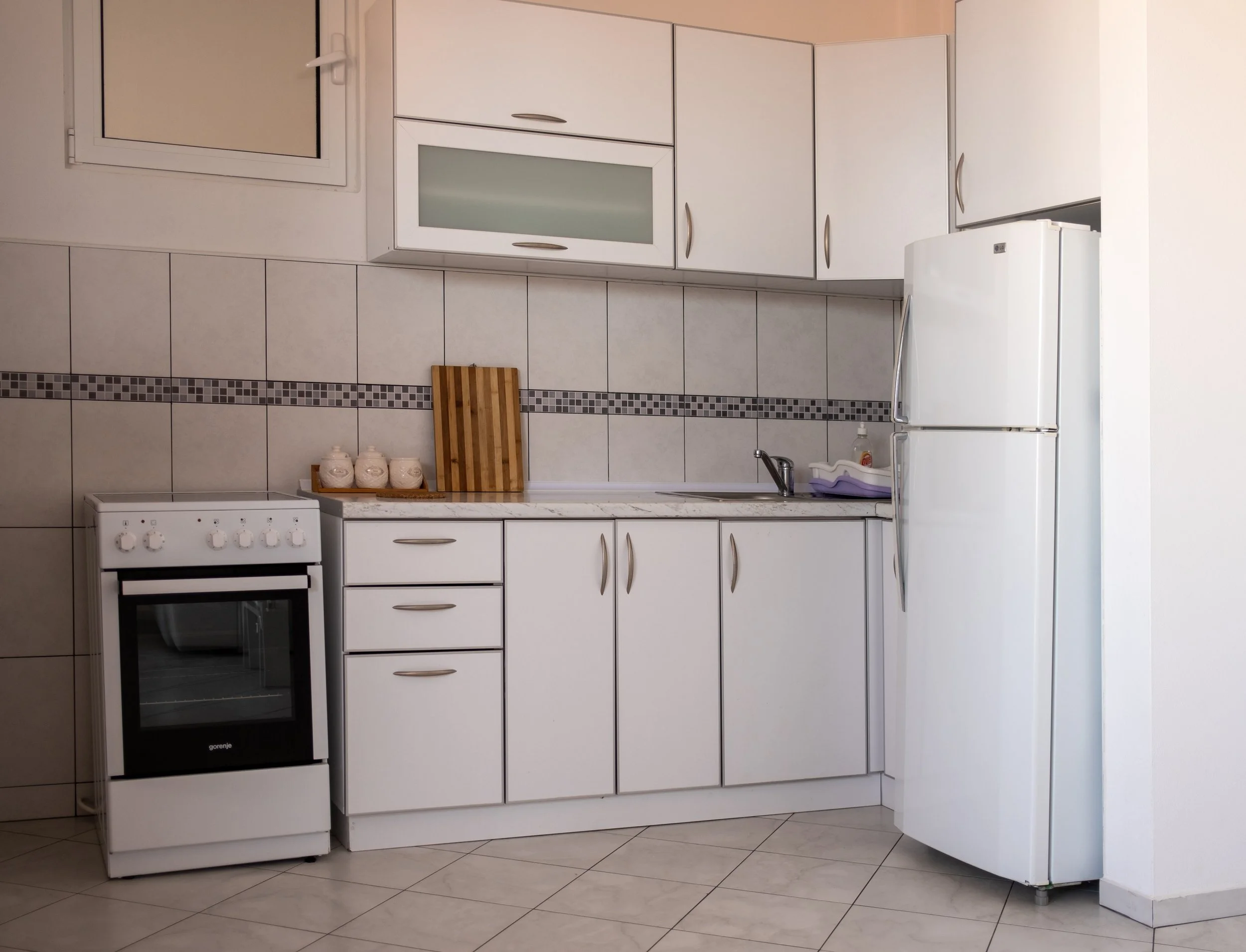 A kitchen with white cabinets, a white stove, a white refrigerator, a sink, tiled walls, and a small window.