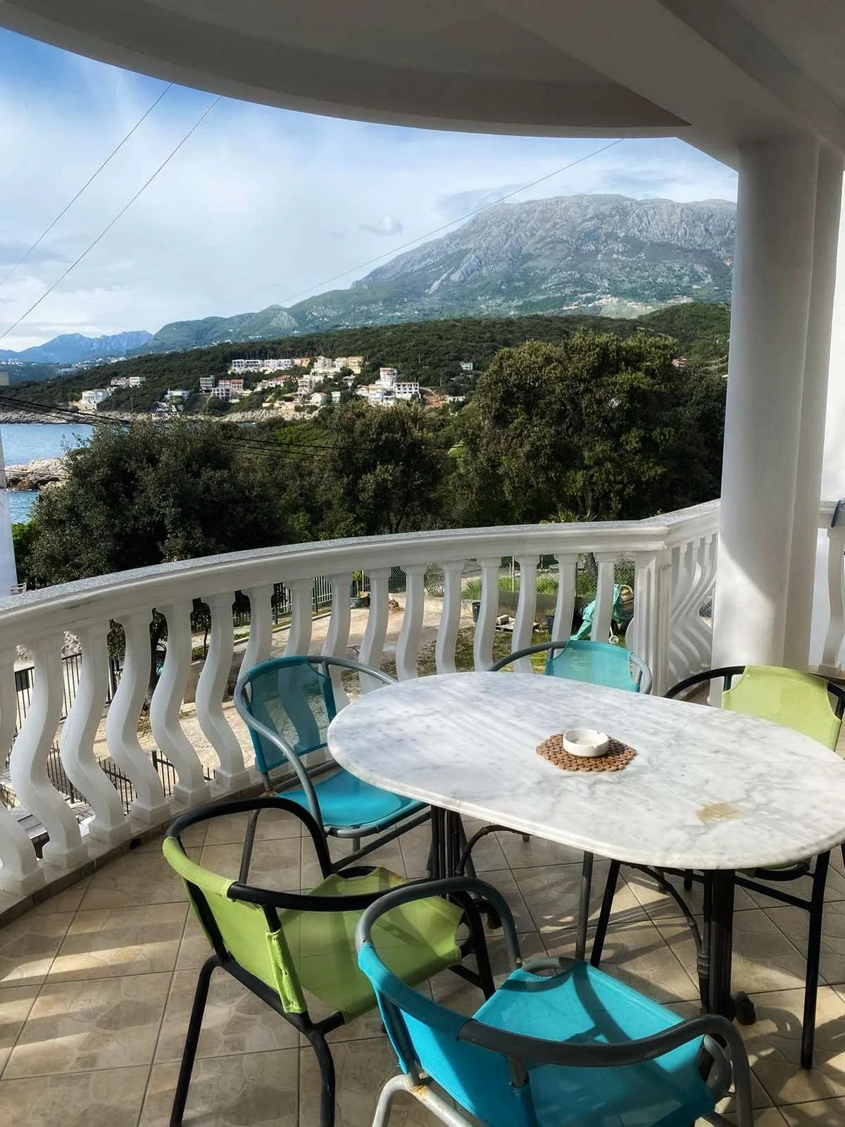 Balcony with white railing, marble table, and colorful chairs overlooking lush green landscape, trees, mountains, and partially cloudy sky.