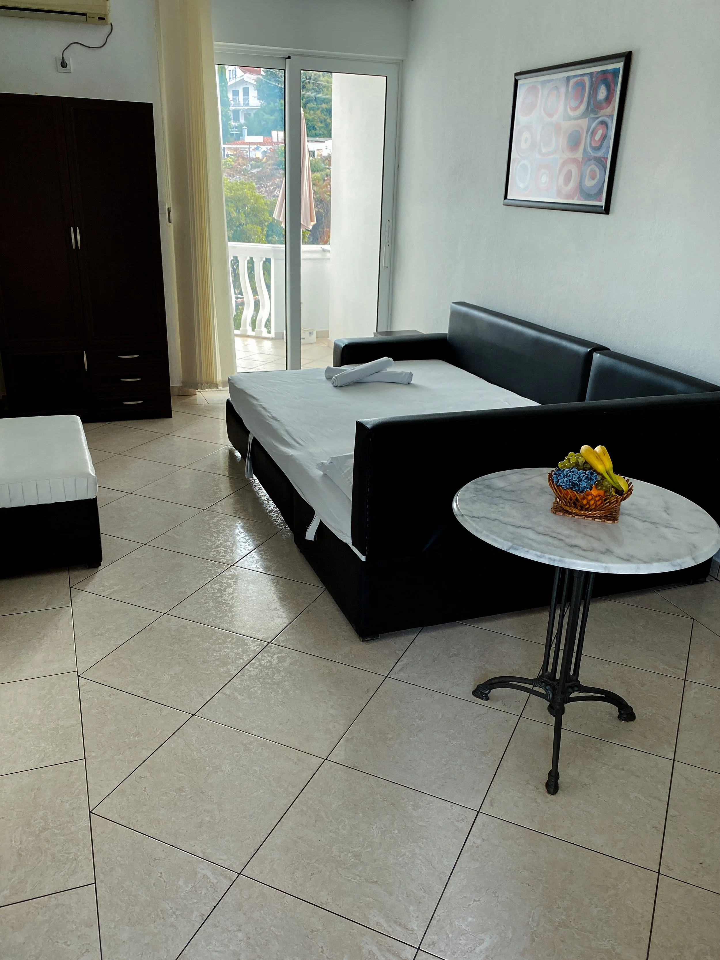 Interior of a living room with a sliding glass door leading to a balcony, a black leather sofa with white sheets, a small round marble-top table with a fruit basket, and a picture hanging on the wall.