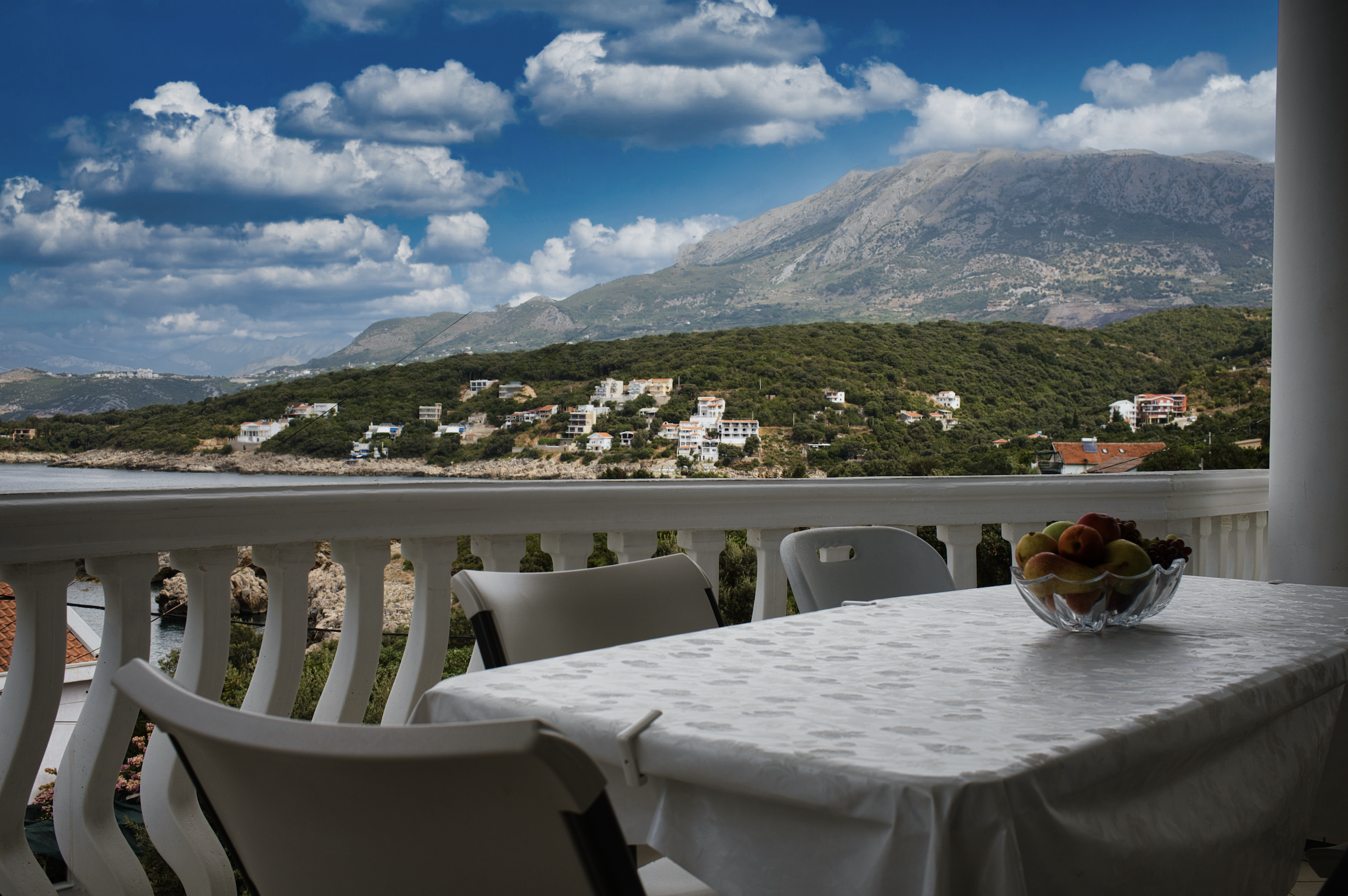View from a balcony showing a mountain landscape with scattered houses, white clouds in a blue sky, and a table with a bowl of fruit on a balcony with white chairs.