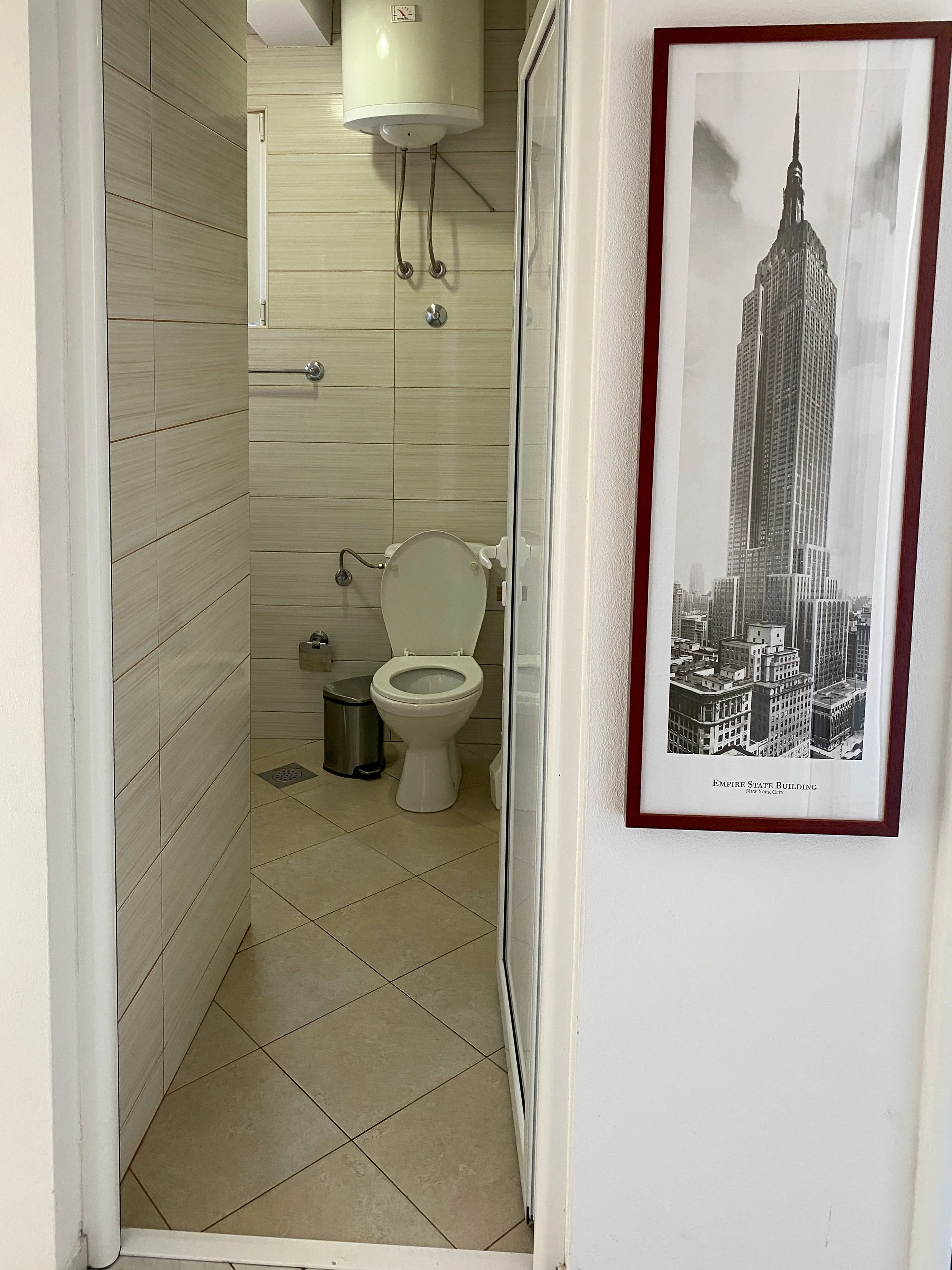 Small bathroom with tiled walls and floor, toilet with open lid, trash can, and water heater on the wall. Framed black and white picture of the Empire State Building hangs on adjacent wall.