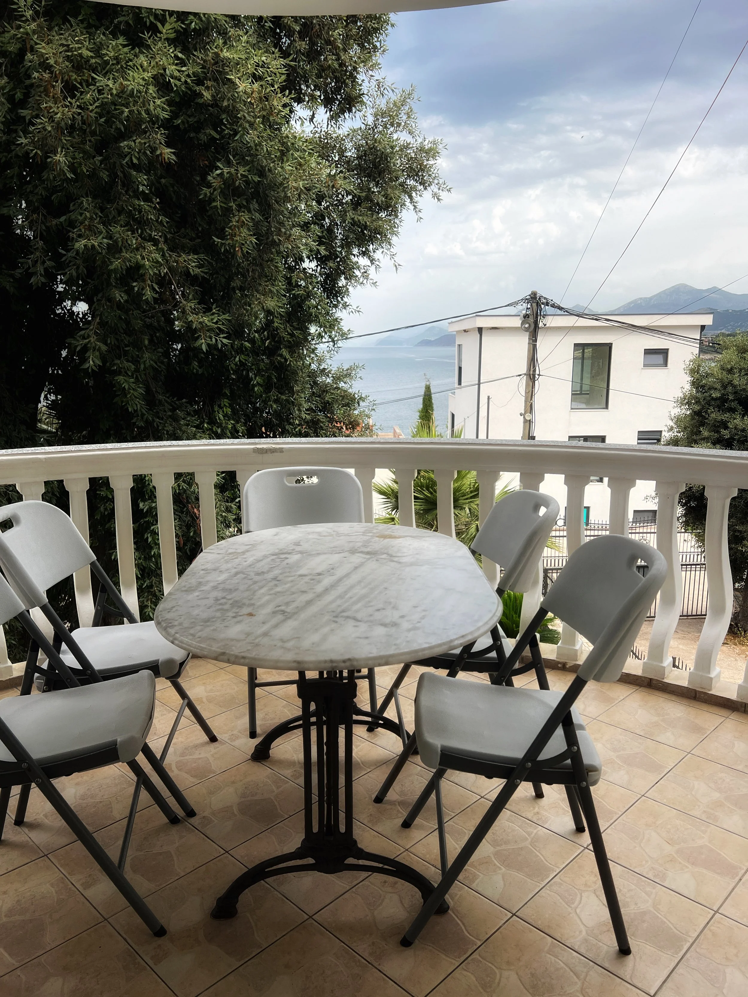 Outdoor balcony with a round marble table and five white chairs with black legs, overlooking a street, trees, a white modern house, power lines, and a view of the sea and mountains in the distance under a partly cloudy sky.