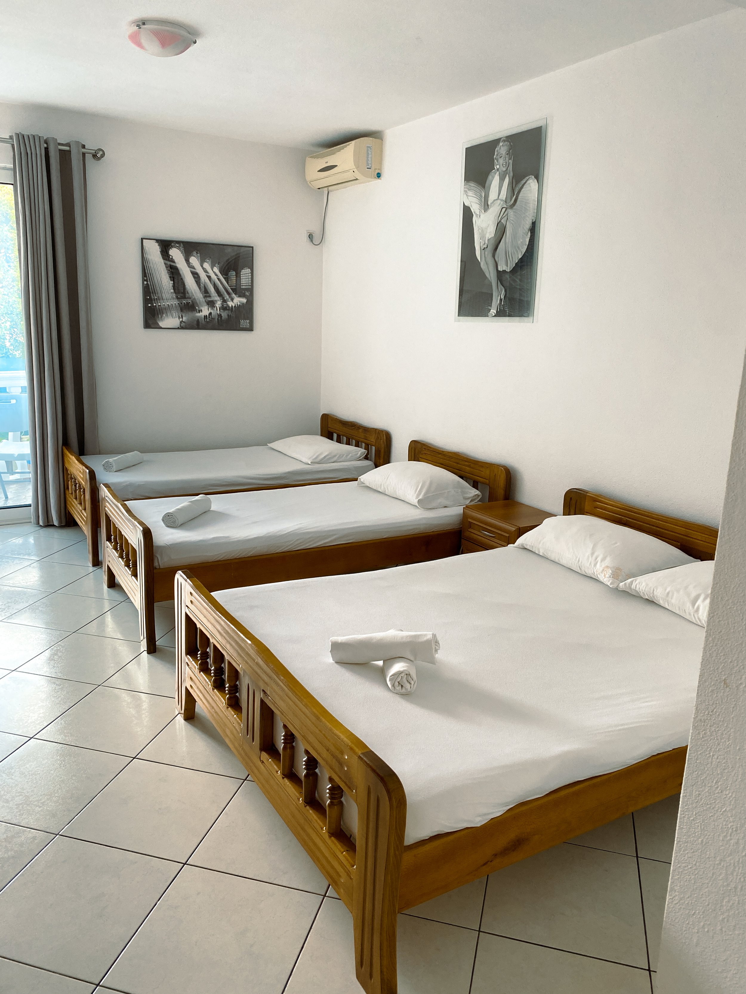 Hotel room with three beds, each with a pillow and a rolled towel, white walls, two framed black and white pictures, a window with curtains, and an air conditioning unit.